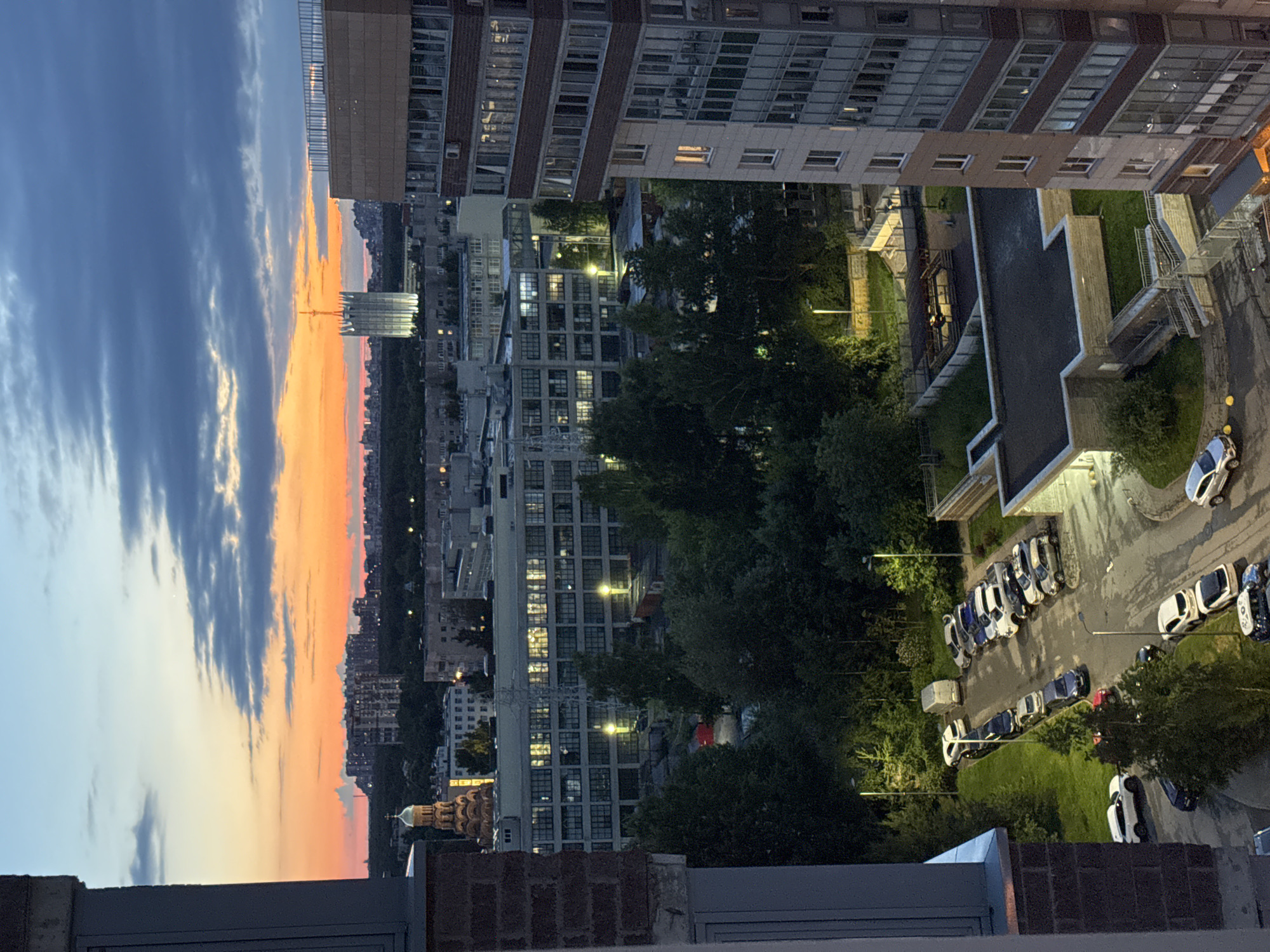 An aerial, elevated, eye-level, medium shot captures a twilight urban landscape in an unknown city in Russia. The sky displays a gradient of colors, from deep blue and grey clouds to vibrant orange and pink hues near the horizon, indicating sunset or sunrise. In the distance, a cluster of apartment buildings forms a skyline, punctuated by a distinctive tall, white, modern building with a pointed top, possibly a landmark or governmental building. Closer to the foreground, a large, multi-story brick and concrete building dominates the right side of the frame. To the left, another brick building is partially visible. Below these structures, a street is lined with parked cars and a few streetlights illuminate the asphalt. Trees are scattered throughout the scene, with denser foliage visible in the mid-ground. The overall impression is one of a residential area as evening sets in.