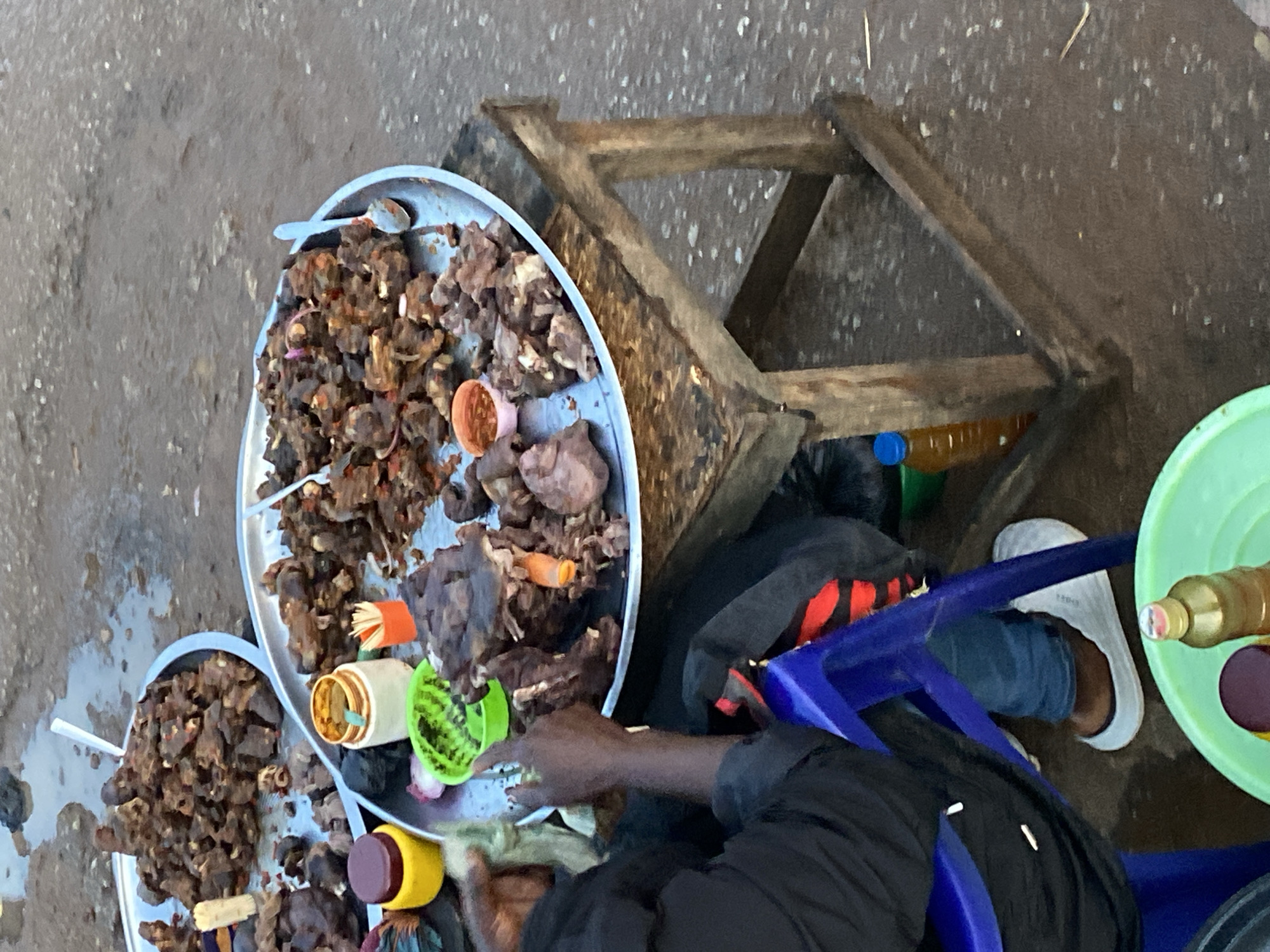 In Bukuru, Nigeria, a vendor displays various meat dishes on metal trays. The dishes are arranged on a wooden table, with some placed on the ground. A person, likely the vendor, is seated on a blue plastic chair, wearing a black jacket and blue jeans, and appears to be interacting with the food. Several small bowls and containers, some holding spices or condiments, are visible on the trays. The ground is wet and appears to be a dirt or unpaved surface, suggesting an outdoor market setting. Bottles of liquids, possibly drinks or oils, are also present.