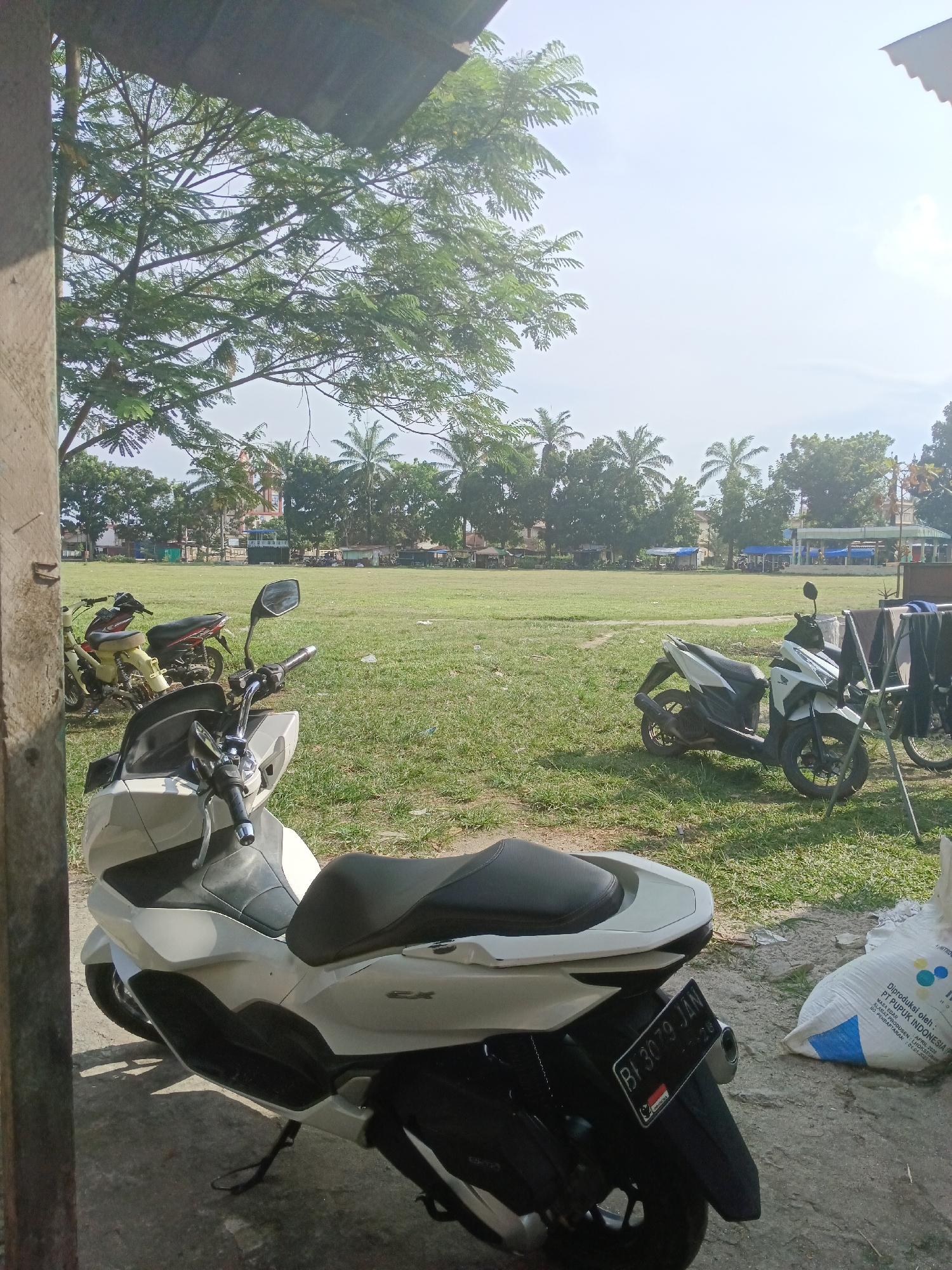 This is a medium-shot, eye-level, outdoor photograph of a paved area and a grassy field under a bright, partly cloudy sky. In the foreground, a white and black scooter is parked, facing left. The scooter has a black seat and a license plate reading "BI 3079 JAN". To the right of the scooter, a large, white bag with blue markings is on the ground. Behind the scooter, there are two other motorcycles parked on the grass. The grassy field extends into the midground, where several palm trees and other trees are visible. In the background, there are buildings, some with blue awnings, and a structure that appears to be a small stadium or grandstand. A clothesline with dark-colored clothes is to the right of the white scooter. The scene appears to be in Aek Kanopan, Indonesia, likely near a public space such as a sports field or open market area.