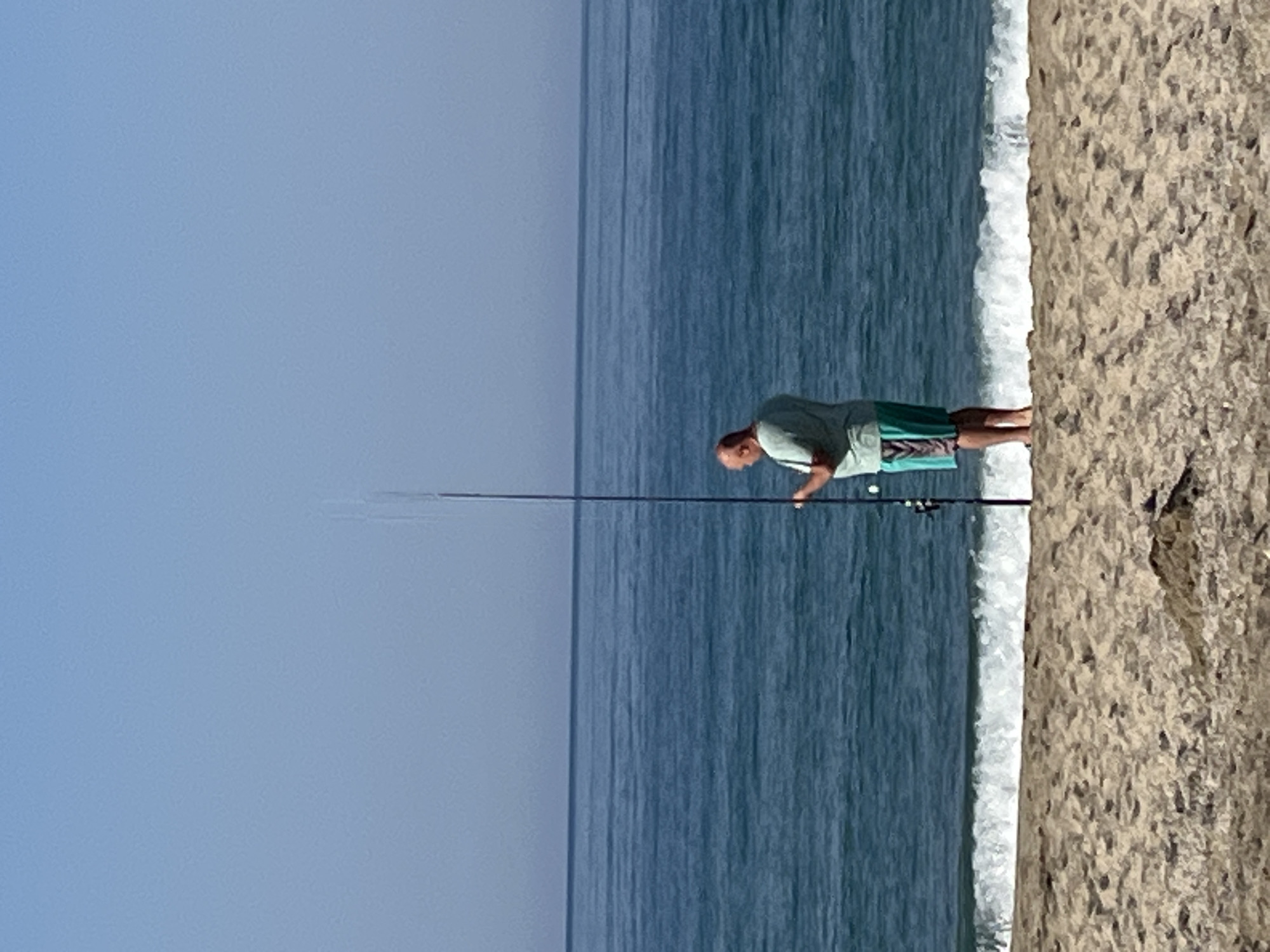 A man is fishing on a beach. He is standing near the shoreline, holding a fishing rod. The scene is likely near Alanya, Turkey, given the prompt's information. The environment consists of the sky, the ocean, a sandy beach, and the fisherman. A wave is breaking along the shore.