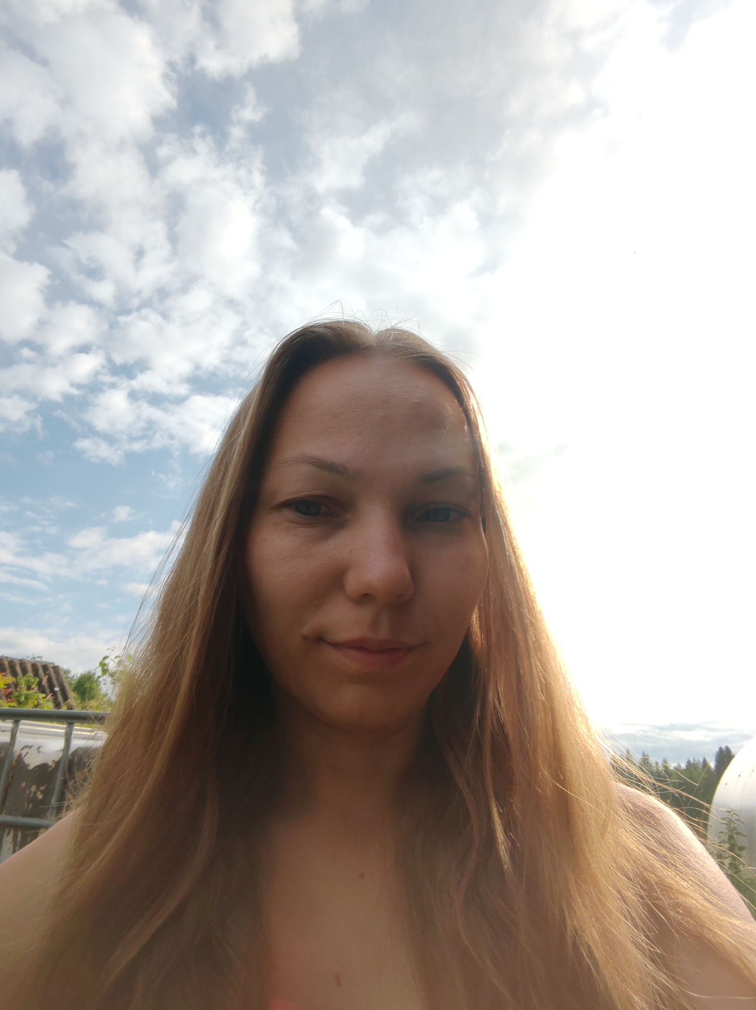 The image is a close-up selfie of a woman with long, blonde hair. She is outdoors with a bright sky and clouds in the background. The sun is shining, creating a lens flare in the upper right portion of the image. The woman is looking at the camera with a neutral expression. The lower part of the image shows her shoulders and chest. There are some trees and a white dome-shaped structure visible in the background, suggesting a rural or semi-rural setting. There are no visible landmarks that would indicate the specific city of Чухлэм, Russia.