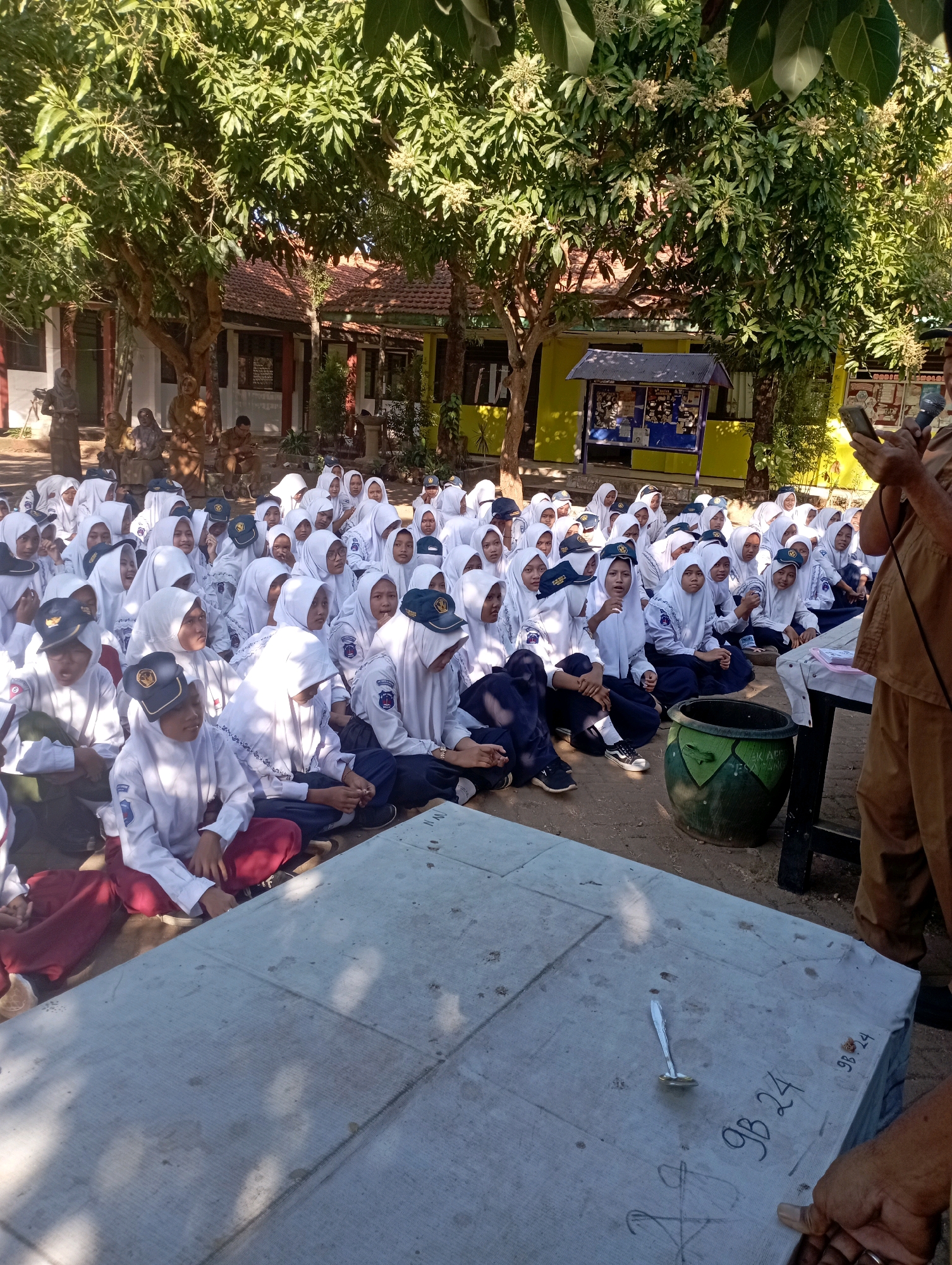 In the city of Banyuurip, Indonesia, under the dappled shade of trees, a group of young students wearing white uniforms and headscarves sit on the ground, facing a table covered with a grey cloth. The students appear to be attending an outdoor class or event. On the table, a small ladle rests on the cloth, which is also marked with handwritten numbers and symbols. In the foreground, a person in a brown uniform stands, holding a mobile phone and a microphone, seemingly addressing the students. Behind the students, a yellow building with a blue bulletin board is visible, suggesting the location is likely a school. The scene is bathed in sunlight, indicating it is daytime.