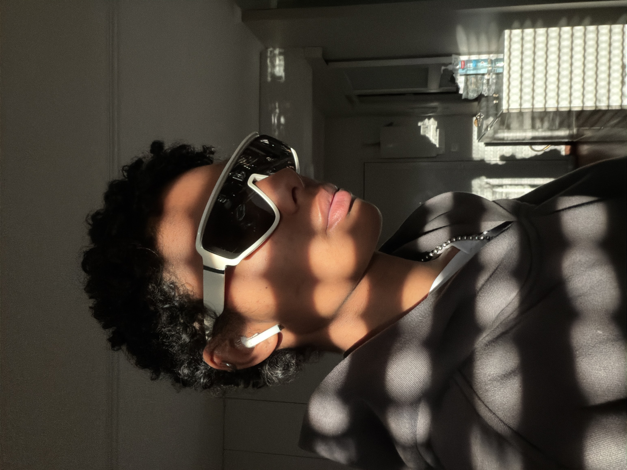 A person with dark curly hair is wearing white-framed sunglasses and a gray hooded sweatshirt. The person's head is tilted back, and they have an earbud in their ear. The light is casting shadows on the person and the background. The background appears to be inside a building, possibly a home, with a refrigerator in the upper right corner of the frame. The location in Champs-sur-Marne, France, cannot be determined from the image.