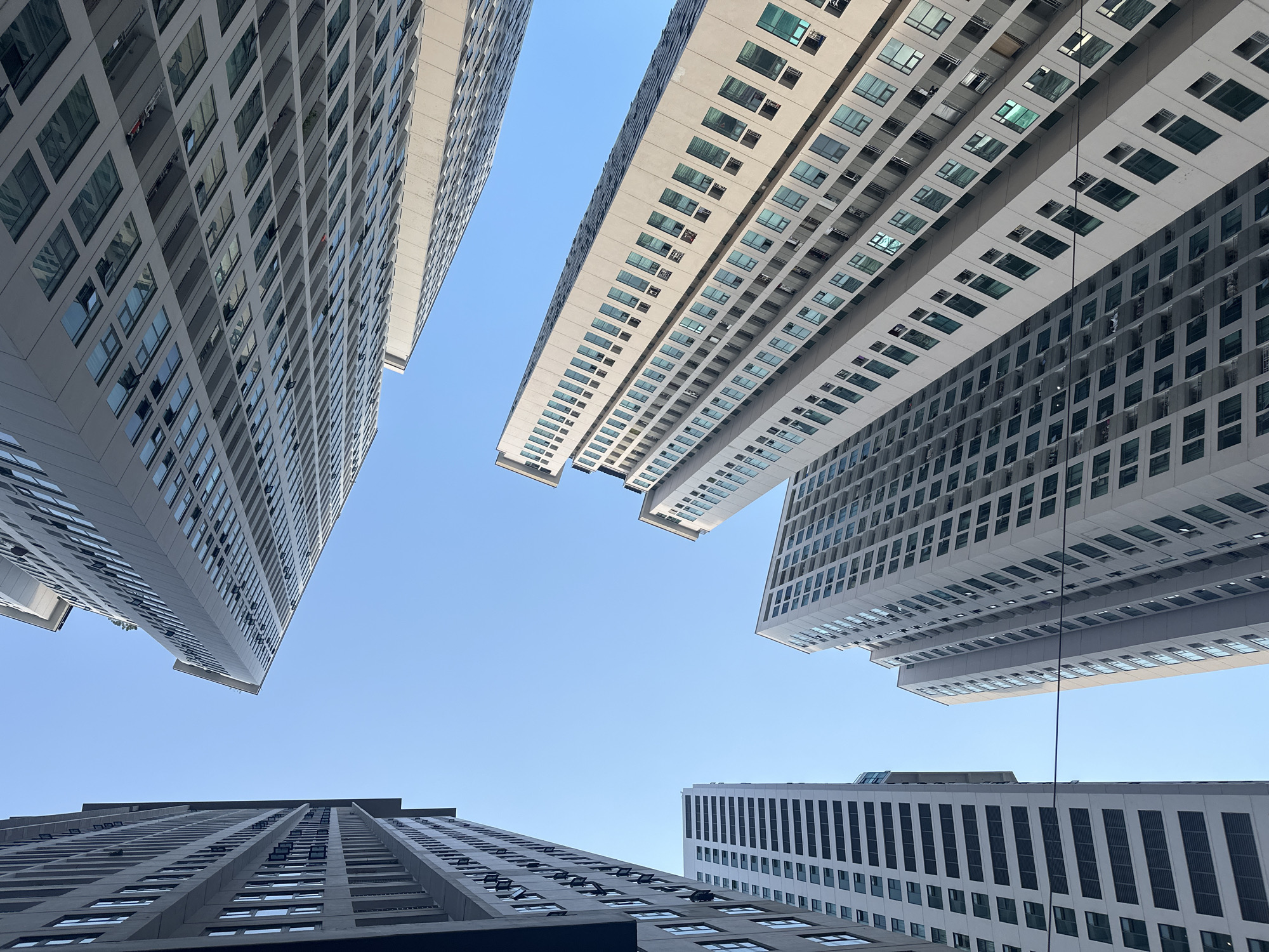The image shows a low-angle view of several tall, modern buildings against a clear blue sky. The buildings appear to be located in a densely built urban area, likely a city center. Their exteriors are primarily gray and white, with rows of windows running vertically along their facades. The buildings are positioned at different angles, creating a dynamic composition. The overall impression is one of urban density and architectural height. The image was taken in the Bac Nha Trang Ward, Vietnam.