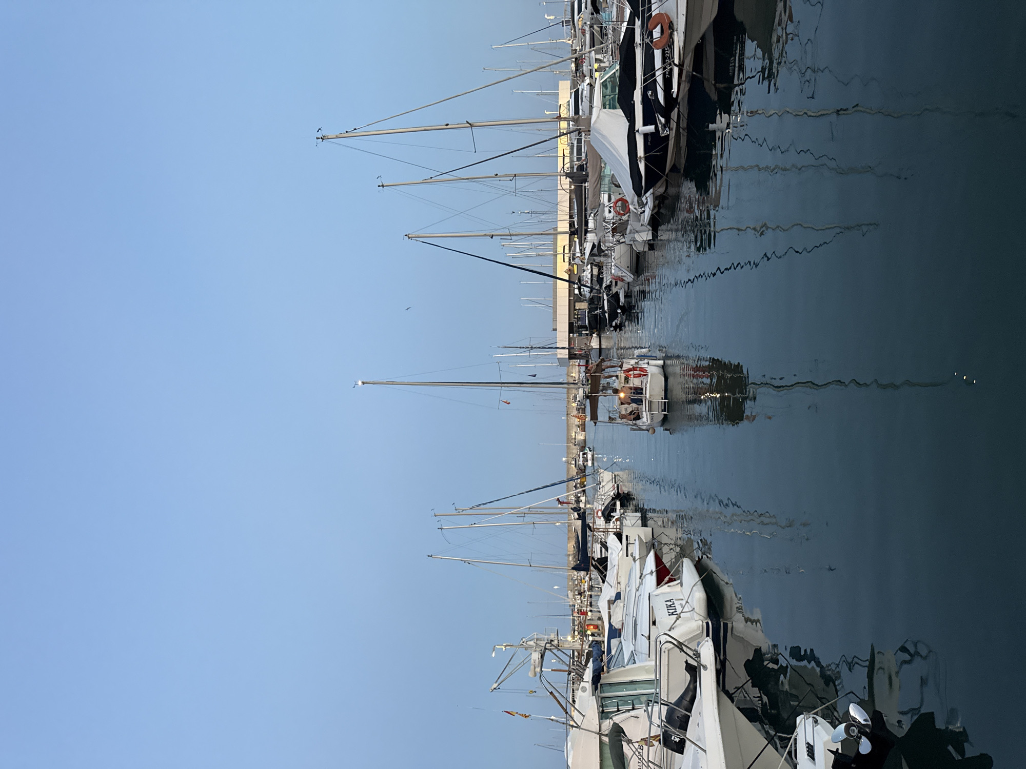 This is a vertical, medium-shot photograph of the Blanes marina in Blanes, Spain, taken at dusk. The sky is a clear, pale blue. The foreground is dominated by a calm, dark body of water reflecting the masts and hulls of numerous moored boats. In the mid-ground, a variety of sailboats and motorboats are docked side-by-side. One sailboat in the center of the frame has several people visible on its deck, some of whom appear to be seated. The boats are of various sizes and designs, with some showing covered hulls. In the background, a modern building with a canopy structure is visible, lit from within. A bridge spans the water further in the distance. Several lights are visible on the boats and along the marina infrastructure. The overall impression is one of a peaceful harbor at the end of the day.