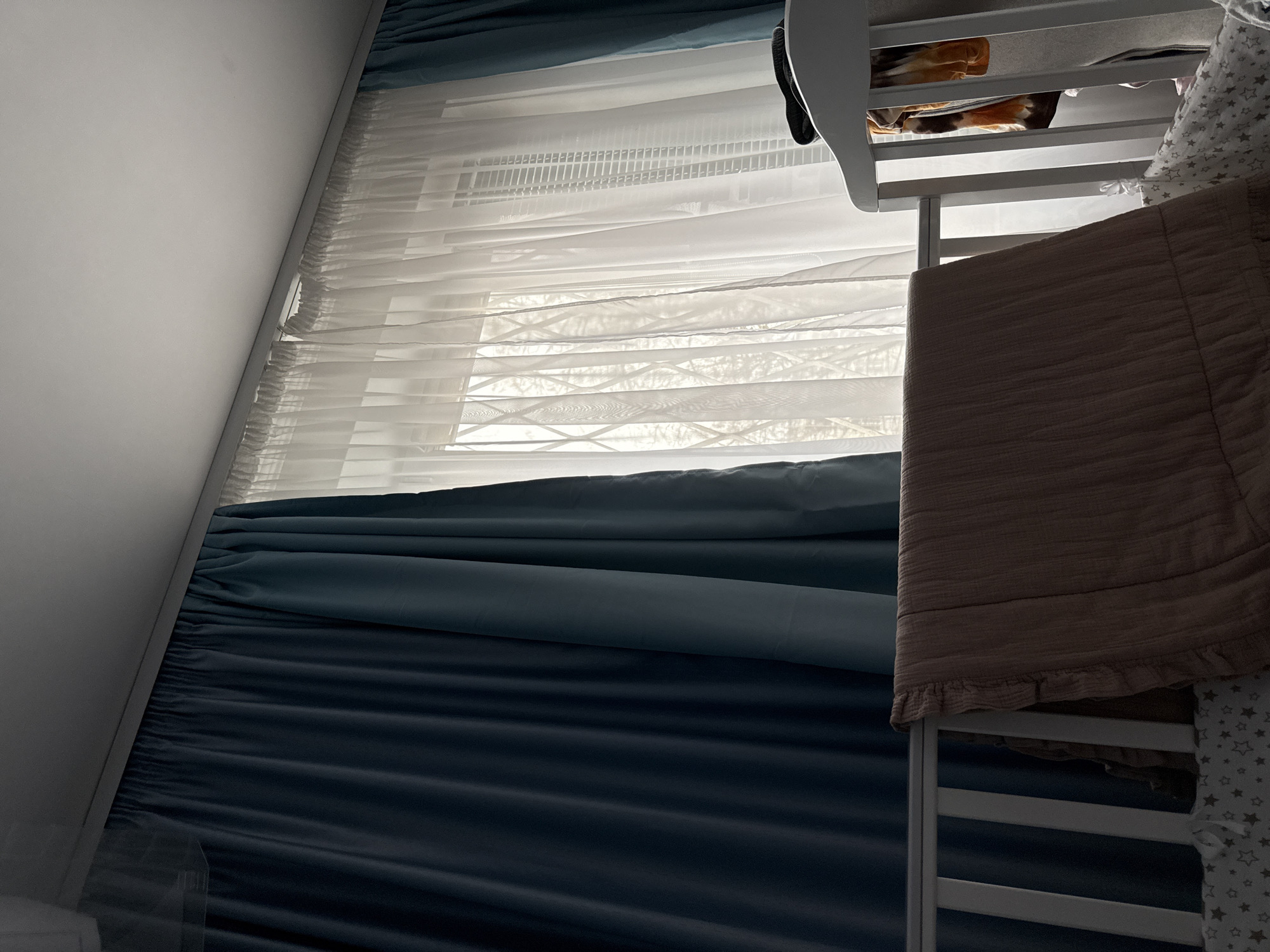 The image shows a corner of a room with a window covered by sheer white curtains and heavier, blue curtains. A white crib with a brown blanket and star-patterned bedding is in the foreground. The room appears to be indoors, with a white wall partially visible. The location is not specified in the image.