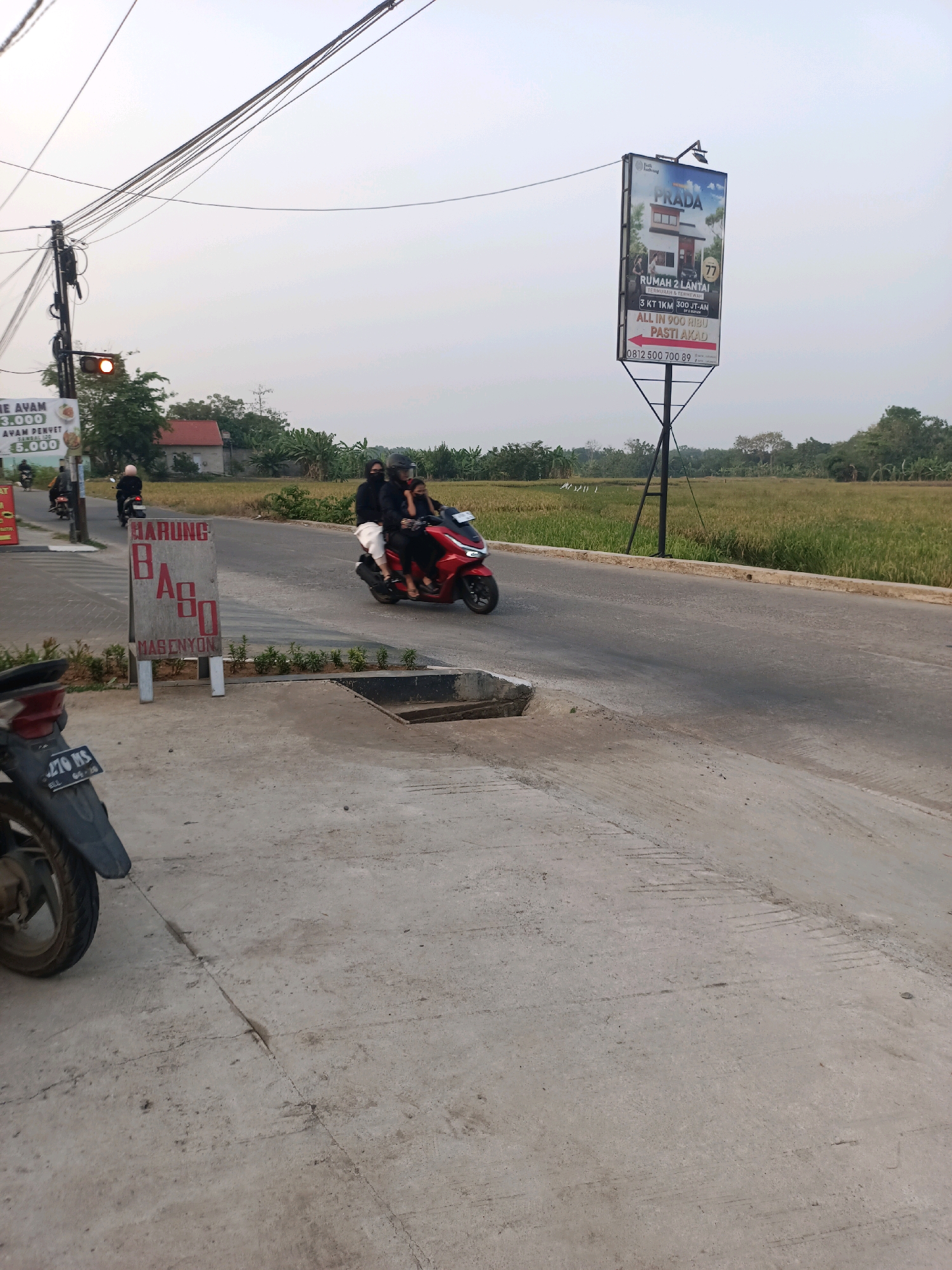 The image depicts a roadside scene in Karawang, Indonesia. In the foreground, a concrete pavement area is visible, with a damaged section near a drainage grate. Part of a parked motorcycle, with license plate "270 MS," is on the left side. A wooden sign for "BARUNG BASO MASENYON" stands on the pavement, with "BARUNG" and "MASENYON" written vertically and "BASO" written horizontally.
In the midground, a red motorcycle with two passengers, both wearing dark clothing and masks, travels along a paved road. Behind them, another motorcycle with a single rider, also in dark clothing, is seen further down the road. On the right side of the road, a large billboard advertising "PRADA" and a housing complex is mounted on a pole. The billboard features an image of a two-story house and text indicating "RUMAH 2 LANTAI, TERMURAH & TERMEGAH" (2-story house, cheapest & most luxurious) with details about land and building size, and contact information.
The background consists of a rice field with golden stalks, suggesting it is harvest season. Scattered trees and some buildings are visible beyond the field. Power lines stretch across the sky, and a traffic light with an orange signal is positioned above the road. The sky is overcast, indicating either early morning or late afternoon. The overall impression is of a rural or suburban area along a main road.