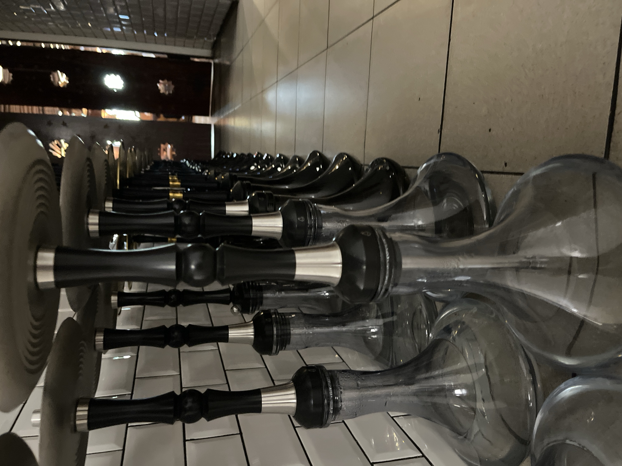 A row of identical shisha pipes stands on a tiled floor in what appears to be a commercial establishment. The shisha pipes have black stems with silver accents and clear glass bases. They are arranged in a line, receding into the background. The environment features a tiled wall behind the shisha pipes and a tiled floor in the foreground and middle ground. In the background, there is a doorway with decorative wooden elements and natural light filtering through. There are no people visible in the image. The lighting suggests an indoor setting with artificial and natural light sources. The perspective is low-angle, looking up at the shisha pipes. The scene is clean and orderly.