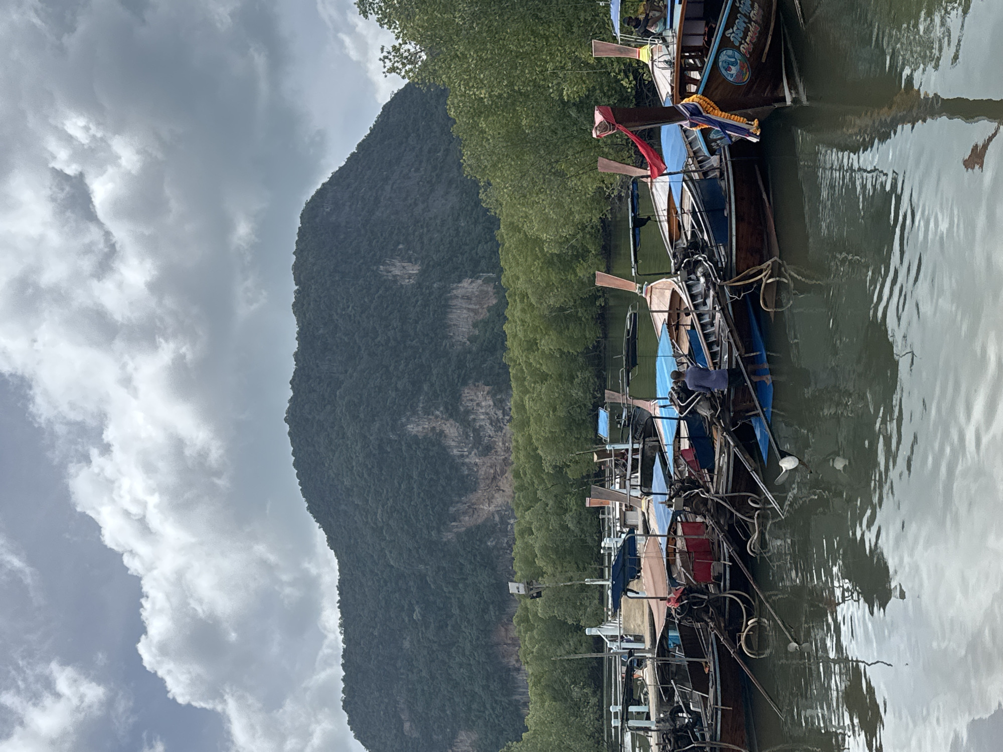 The image depicts a scene at a pier in Ban Hin Rom, Thailand, featuring a row of traditional longtail boats docked in the water. The boats, primarily made of wood, are varied in color with blue and brown canopies providing shade. Some boats are adorned with decorative ribbons and tassels. In the foreground, the water reflects the boats and the overcast sky. A person in a blue shirt is visible standing on one of the boats, appearing to be attending to it. In the background, a dense green forest lines the water's edge, with a large, tree-covered mountain rising behind it. The sky is filled with dramatic, grey clouds, suggesting an impending change in weather. The overall atmosphere is one of a working harbor, with the natural landscape providing a dramatic backdrop.