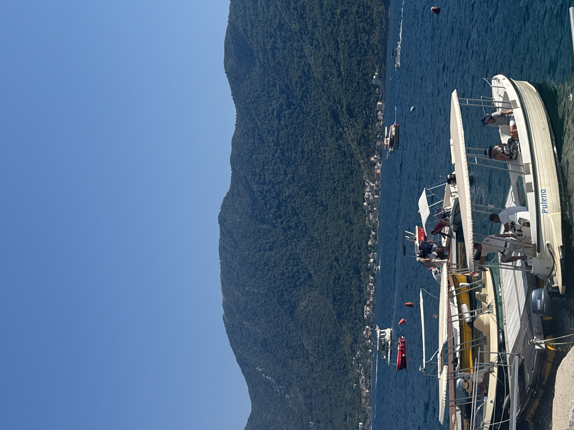 In the city of Perast, Montenegro, a cluster of small boats, some with canopies, are docked at a pier on a bright, sunny day. The deep blue water of the bay is calm, with a few other boats in the distance. In the background, a large, verdant, tree-covered mountain slopes down towards the water, dotted with the red-tiled roofs of houses and buildings, indicative of the town's architecture. On the pier, several people are visible, some standing and some seated in the boats. One boat in the foreground has the name "Pulena" written on its hull. Another boat has a Yamaha outboard motor. The scene evokes a sense of leisure and coastal tourism.