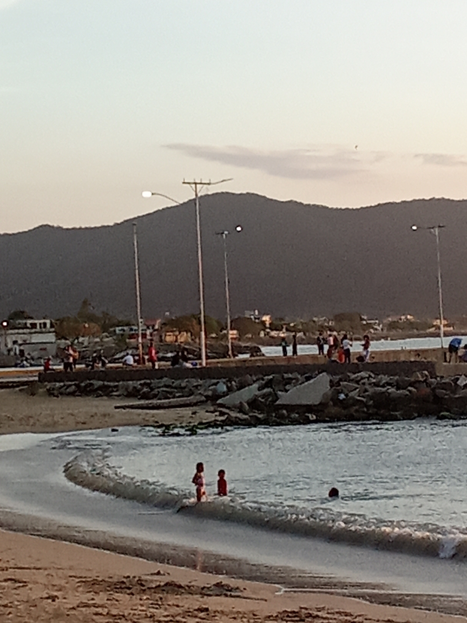 The image displays a coastal environment in Juan Griego, Venezuela, under twilight conditions. The foreground consists of a sandy beach transitioning into calm, shallow water where three individuals, appearing to be children, are wading; two are standing upright, and one is partially submerged. The midground is dominated by a concrete pier or jetty that extends into the water from the left, supported by a base of large, irregular rocks. Numerous indistinct human figures are distributed along the pier. Several tall streetlights with illuminated fixtures are positioned on or near the pier and shoreline. In the background, a developed coastal area with buildings and vegetation is visible, leading to a prominent, dark mountain range under a pale, largely clear sky. A single, small, dark, unidentifiable object is present in the upper right quadrant of the sky.