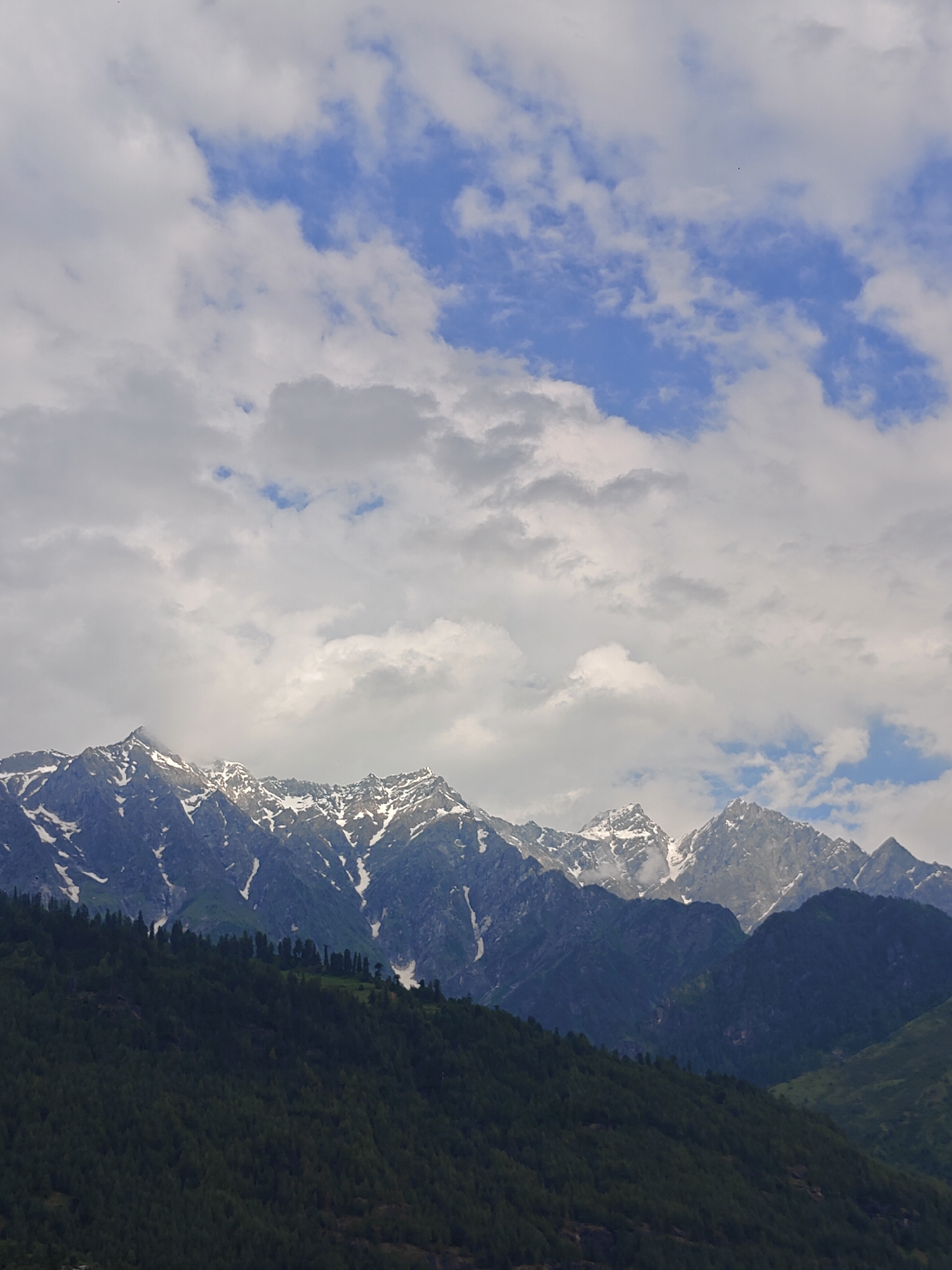 The image depicts a majestic mountain range, likely in the vicinity of Vashisht, India, given the description. The peaks are snow-capped, contrasting sharply with the dark green, densely forested slopes that rise from the foreground. The sky above is a dynamic canvas of white, fluffy clouds interspersed with patches of bright blue. The overall scene is one of natural grandeur and serene beauty, characteristic of Himalayan landscapes.