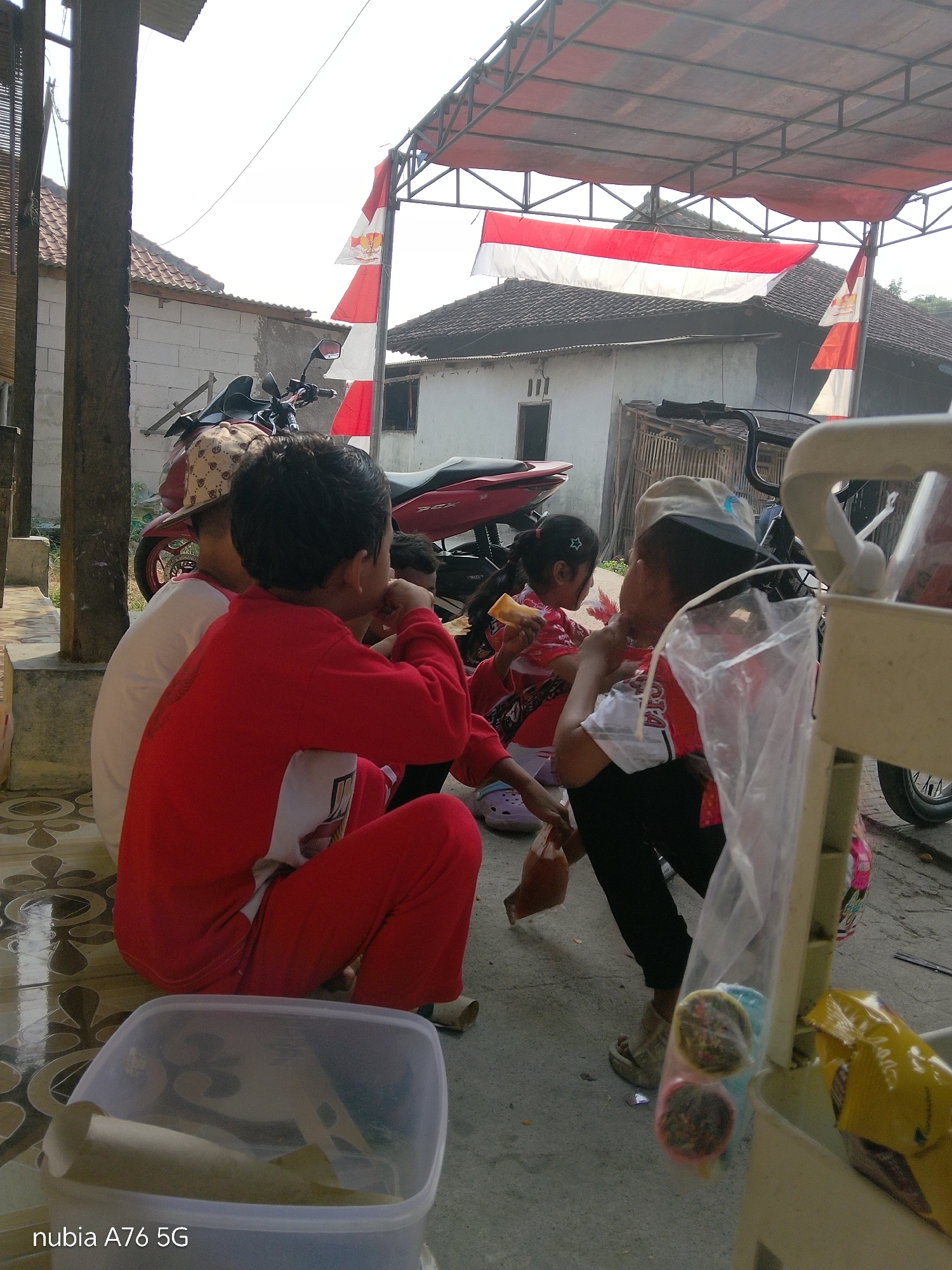 In the city of Binuang, Indonesia, a group of children are gathered outdoors, possibly in a residential yard, with a motorcycle and residential buildings visible in the background. Two children, one in a red shirt and pants and the other wearing a patterned cap and white shirt, are seated on the ground. A third child, a girl in a red and white patterned shirt, is also seated and holding a piece of food. Another child, a boy in a red shirt and black pants, is leaning forward. A food vendor's cart is partially visible on the right, with plastic-wrapped snacks hanging from it. Indonesian flags are displayed on poles and under a canopy, suggesting a national celebration or event. The ground is paved with patterned tiles.