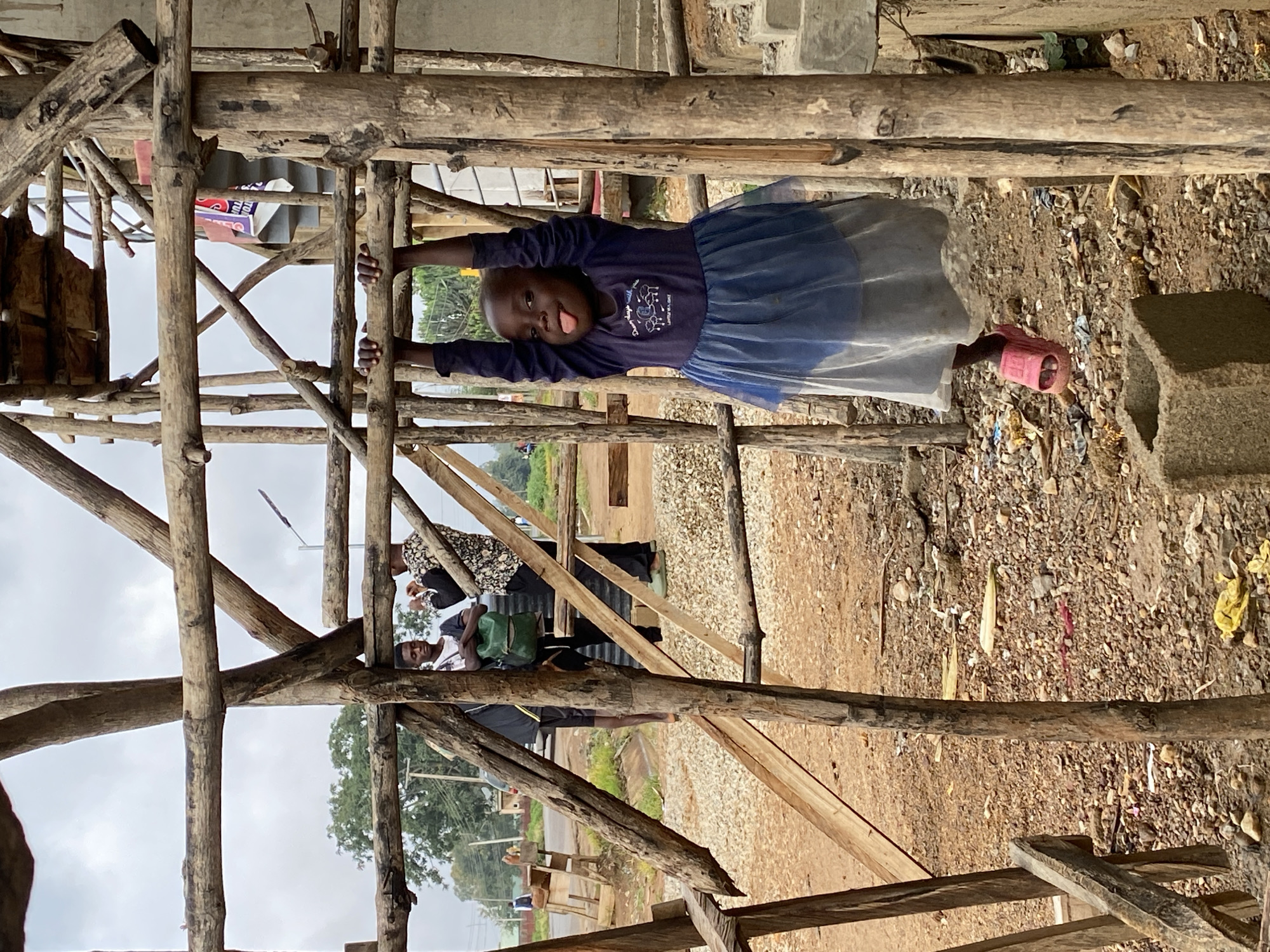 This image, set in Riyom, Nigeria, captures a young child playfully hanging from a rough wooden structure, likely scaffolding or part of an unfinished building. The photograph is oriented vertically, with the child as the central subject.
The child, with dark skin and a bald or closely shaved head, is seen in the middle right of the frame, holding onto two horizontal wooden beams. She wears a dark blue long-sleeved shirt with a white graphic design and partially visible text that reads "Dream big, work hard." A light blue or grey sheer, tutu-style skirt completes her outfit, and a single pink slipper is visible on her right foot. She is looking directly at the camera with a playful expression, sticking out her tongue.
The wooden structure, made from untrimmed logs and branches, dominates the foreground and midground, forming a rustic, crisscrossing framework. Below the child, the ground is unpaved, consisting of dirt, gravel, and small rocks, with some scattered debris. A concrete block is visible on the ground to the right.
In the background, beyond the wooden lattice, several other people are visible. One individual wears a black and white patterned top, and another wears a black t-shirt, shorts, and carries a green backpack, standing on what appears to be a dirt road or path. Green trees and foliage are also visible in the background, set against a grey, overcast sky, indicating it is daytime. The scene suggests a rural or semi-urban environment, possibly an area under construction or with basic infrastructure.