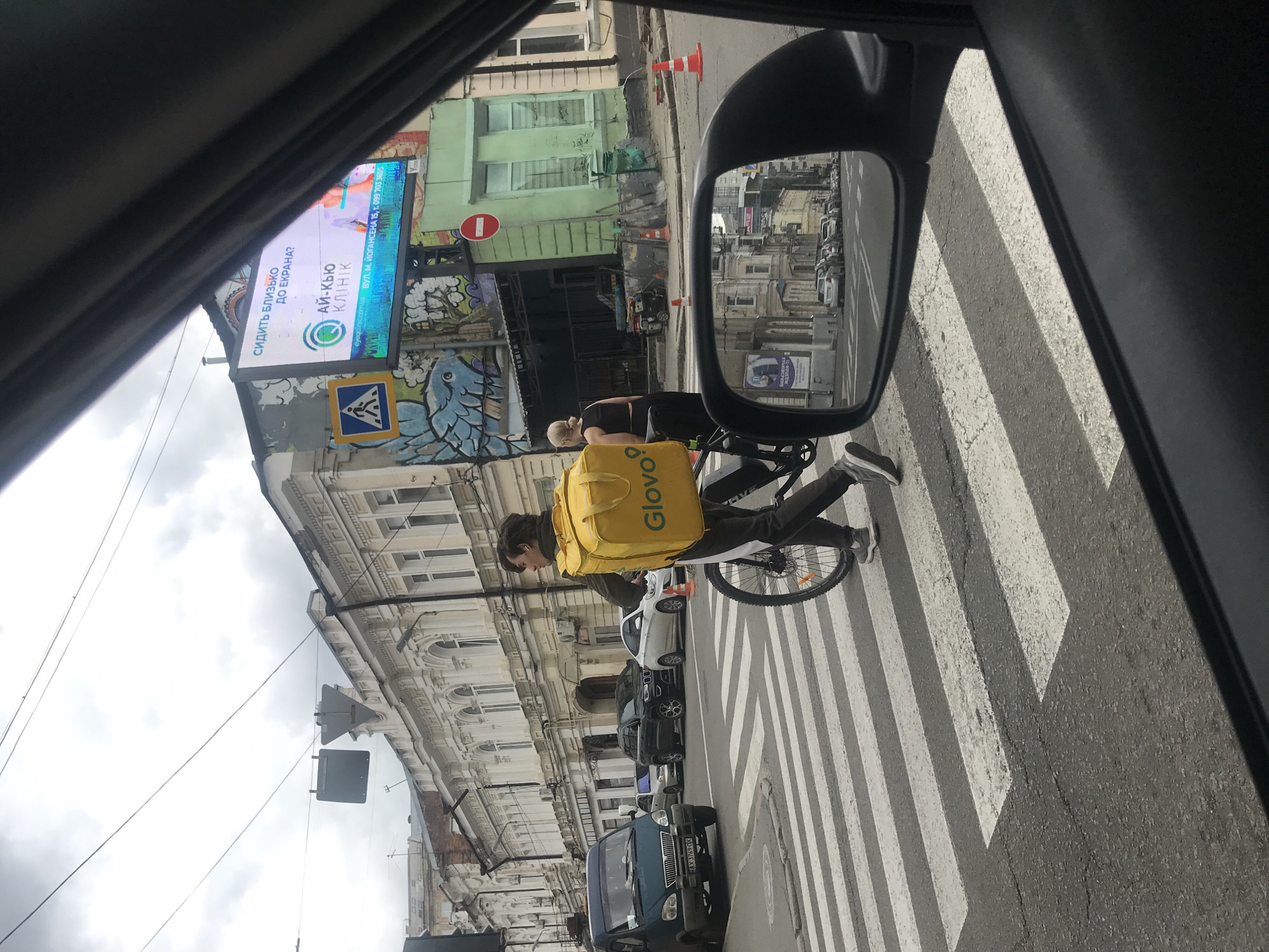 Here's a concise description of the scene:
The image, taken from inside a car, shows a Glovo delivery person riding a bicycle across a pedestrian crosswalk in Kharkiv, Ukraine. The cyclist has a bright yellow Glovo delivery bag. Cars are lined up along the street. A large advertising screen displaying information about the "Eye-Q Clinic" is visible on a building in the background. There is a road sign and graffiti on the building, as well as multiple stories of apartments. A car's side mirror is in the shot, showing other buildings and vehicles behind. A vehicle with the license plate AX 3769 OX is visible parked in the foreground.