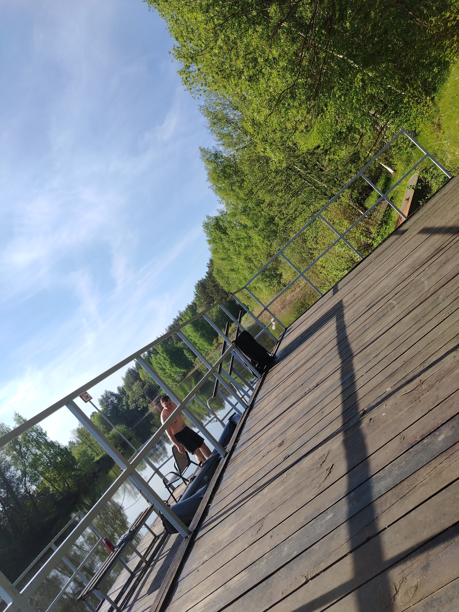 A daytime, angled shot shows a wooden dock extending into a body of water in the city of Stroitel', Russia. A man wearing dark shorts stands on the dock, near a metal railing that encloses the dock's perimeter. Behind the man, a chair and a boat are visible. Trees with green foliage form the background, lining the edge of the water. The sky is blue with scattered clouds. The wooden planks of the dock cast long shadows, indicating sunlight.
