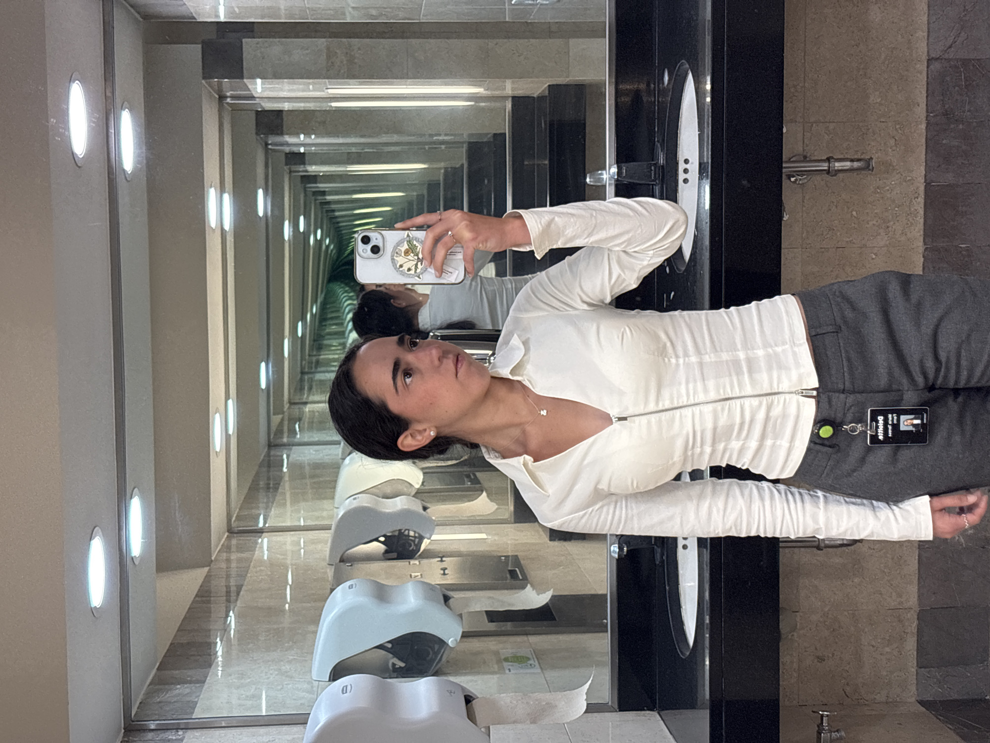 A woman is taking a selfie in a bathroom with an infinity mirror effect. The bathroom is located in Mexico City, Mexico, likely within an office building or upscale public space. The woman is wearing a white long-sleeved top and grey pants. She is holding an iPhone with a patterned case. She has dark hair pulled back. The bathroom has automatic hand dryers and dark countertops with light-colored sinks. A name tag on the woman's pants reads "Laura Deloitte". The mirror creates a seemingly endless hallway with lights. The bathroom appears to be clean and well-maintained.