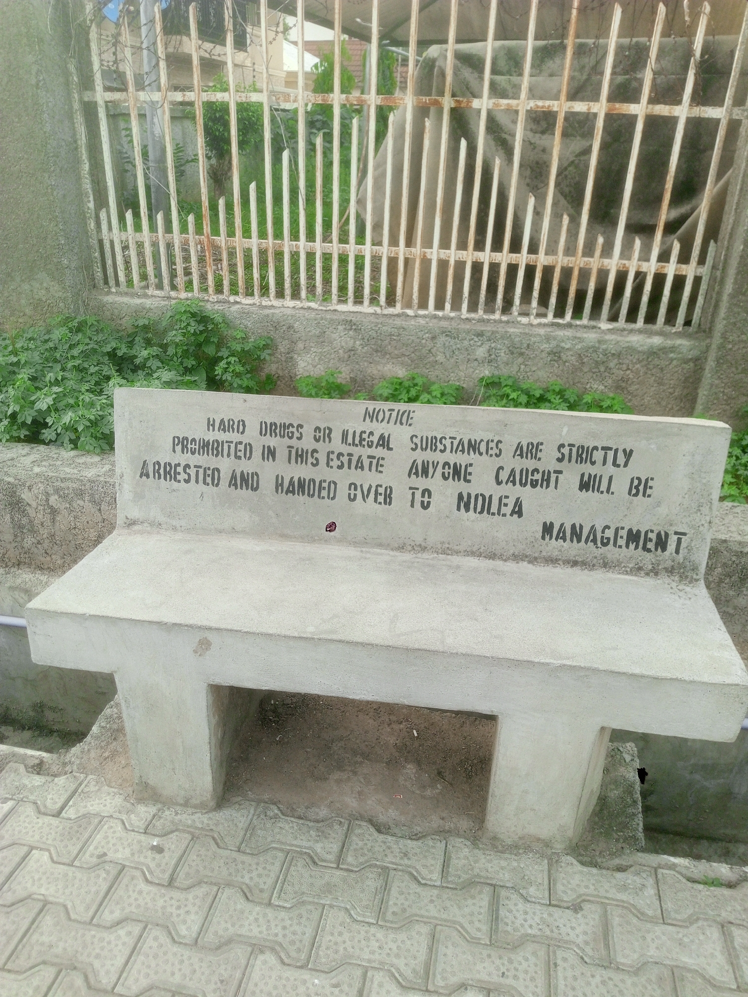 The image captures a static outdoor scene featuring a concrete bench with a prominent warning message inscribed on its backrest. The setting appears to be within or adjacent to an "estate," as indicated by the text, in Abuja, Nigeria.
The primary subject is the sturdy, light grey concrete bench. Its backrest displays stenciled black capital letters with the following notice: "NOTICE HARD DRUGS OR ILLEGAL SUBSTANCES ARE STRICTLY PROHIBITED IN THIS ESTATE ANYONE CAUGHT WILL BE ARRESTED AND HANDED OVER TO NOLEA MANAGEMENT." This explicit message serves as a clear warning against drug-related activities, enforced by Nigeria's National Drug Law Enforcement Agency (NOLEA).
Behind the bench, a white metal fence, possibly a gate or part of a perimeter, shows signs of weathering and minor rust. Beyond the fence, green foliage and bushes are visible, growing against a concrete wall. The ground around the bench is paved with grey interlocking concrete blocks, suggesting a designated public or common area. The lighting indicates it is daytime, possibly with overcast conditions given the diffused light and absence of harsh shadows. There are no people visible, and no specific activity is taking place; the image focuses solely on the warning sign and its immediate surroundings. The scene communicates a message of strict regulation and law enforcement regarding prohibited substances within the estate.