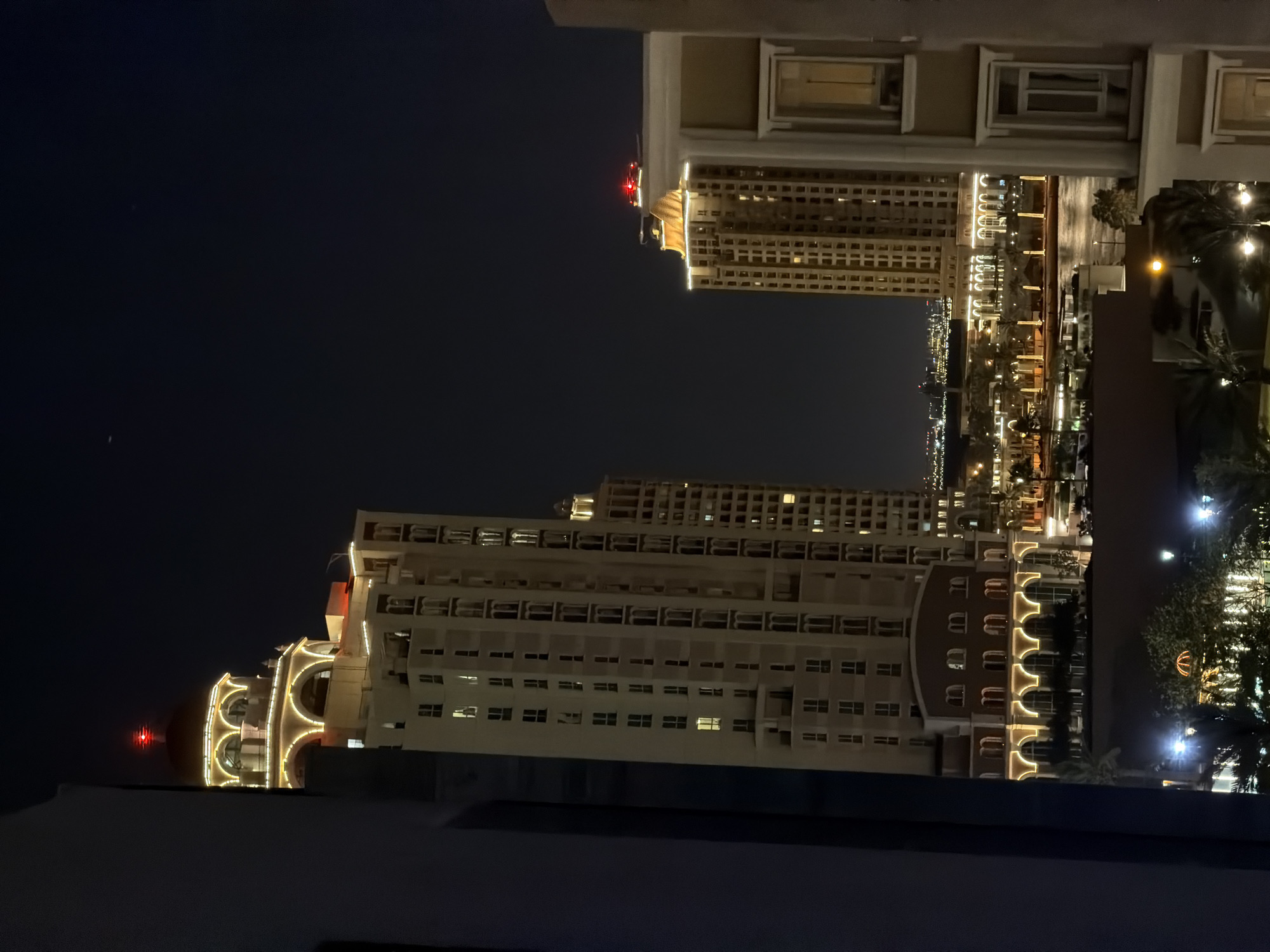 This nighttime image captures an urban waterfront scene, likely within The Pearl-Qatar in Doha, Qatar. The perspective is from a building, with parts of the surrounding structure framing the view on the left and right.
In the mid-ground, two prominently illuminated, multi-story buildings dominate the scene. The taller building on the left features a distinctive dome structure at its apex, which is topped with a bright red aviation obstruction light. Decorative yellow and white string lights outline its many arched windows, balconies, and architectural details, including a series of large arches on its lower levels. Adjacent to it on the right stands another tall, light-colored building with a similar architectural style, also featuring decorative lighting and topped with a red aviation obstruction light.
Between these buildings, a body of water, likely a canal or bay, reflects the artificial lights from the surrounding structures. Along the waterfront, streetlights illuminate palm trees and other landscaping. In the background, a distant city skyline, densely dotted with numerous lights, stretches across the horizon. The sky is dark blue to black, indicating nighttime, with a faint white speck visible in the upper central portion. No individuals are depicted in the image.