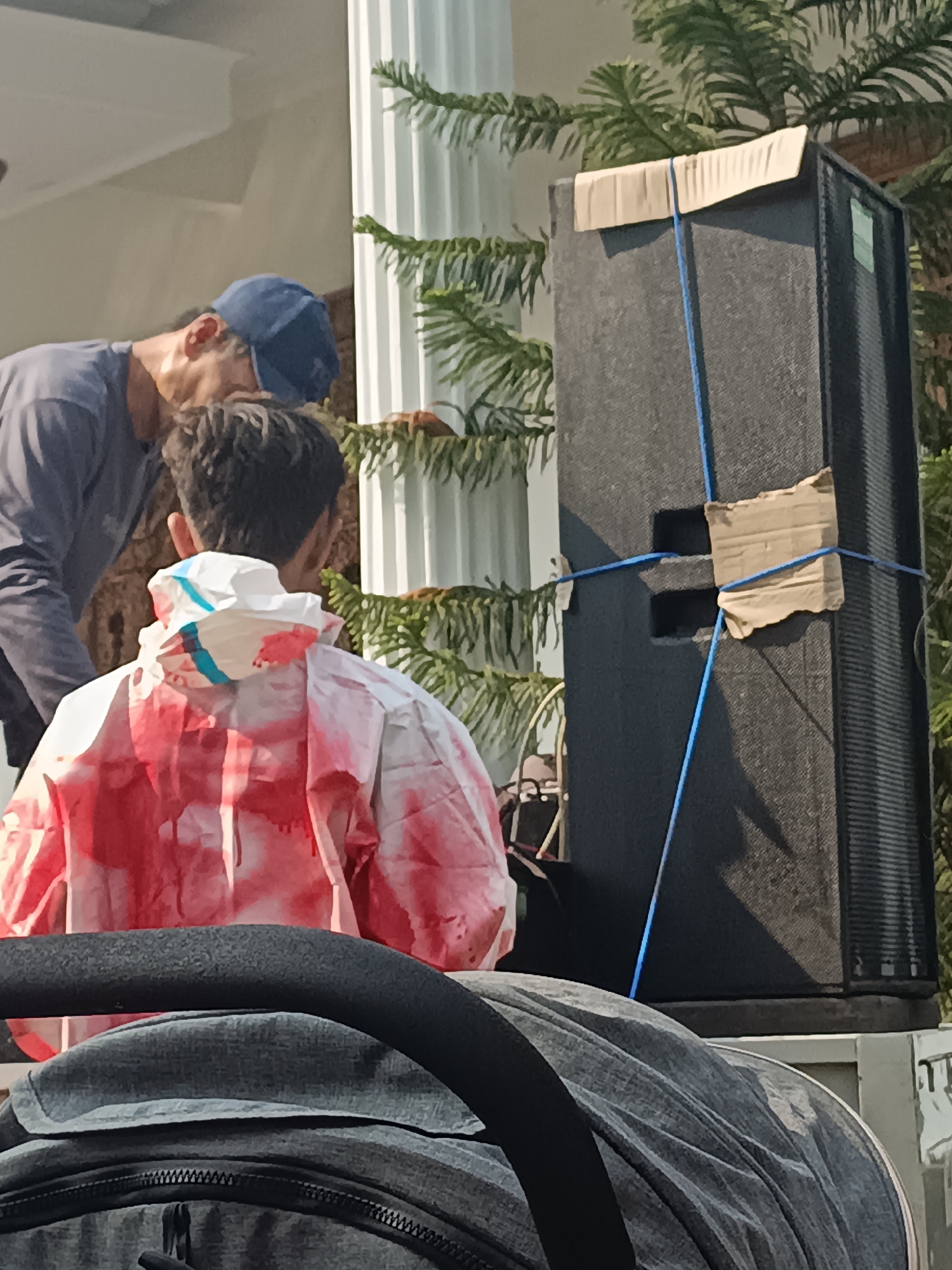 In Sleman, Indonesia, two individuals are observed near a large black speaker. One person, wearing a blue cap and a dark grey long-sleeved shirt, is bent over, appearing to interact with another person. The second individual, facing away from the camera, is wearing a white garment with red splotches, resembling a hazmat suit or a creatively decorated jacket, and is positioned in front of the speaker. The speaker itself is a tall, rectangular unit secured with blue cords and pieces of cardboard. In the foreground, the top of a grey stroller or carrier is visible, partially obscuring the lower portion of the scene. The background includes architectural elements, likely a white column, and lush green foliage, suggesting an outdoor setting with some vegetation.