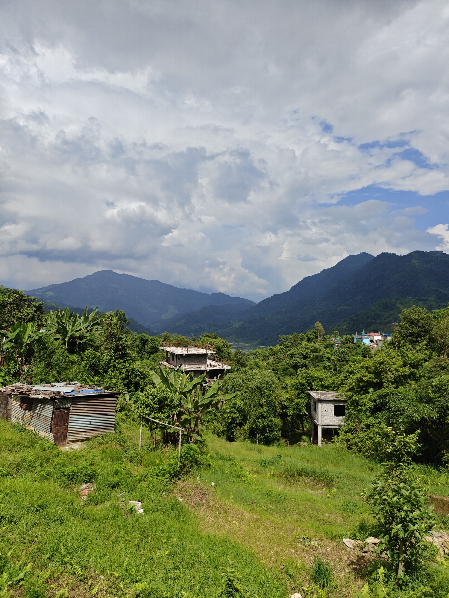 This is a landscape photograph taken in Pokhara, Nepal. The scene depicts a verdant, hilly terrain under a cloudy sky. In the foreground, to the left, is a small, dilapidated structure with a corrugated metal roof and a wooden door. Adjacent to it is a patch of overgrown grass. To the right of this structure, further into the frame, is an unfinished two-story building constructed with concrete blocks, with open window frames. In the middle ground, amidst lush green trees and banana plants, are more structures, including a house with a light blue roof and some other buildings. The background is dominated by rolling, densely forested mountains that recede into the distance, suggesting a valley or a mountain range. The sky is filled with a mix of heavy grey clouds and patches of blue sky. The overall impression is of a rural or semi-rural settlement nestled within a natural, mountainous environment.