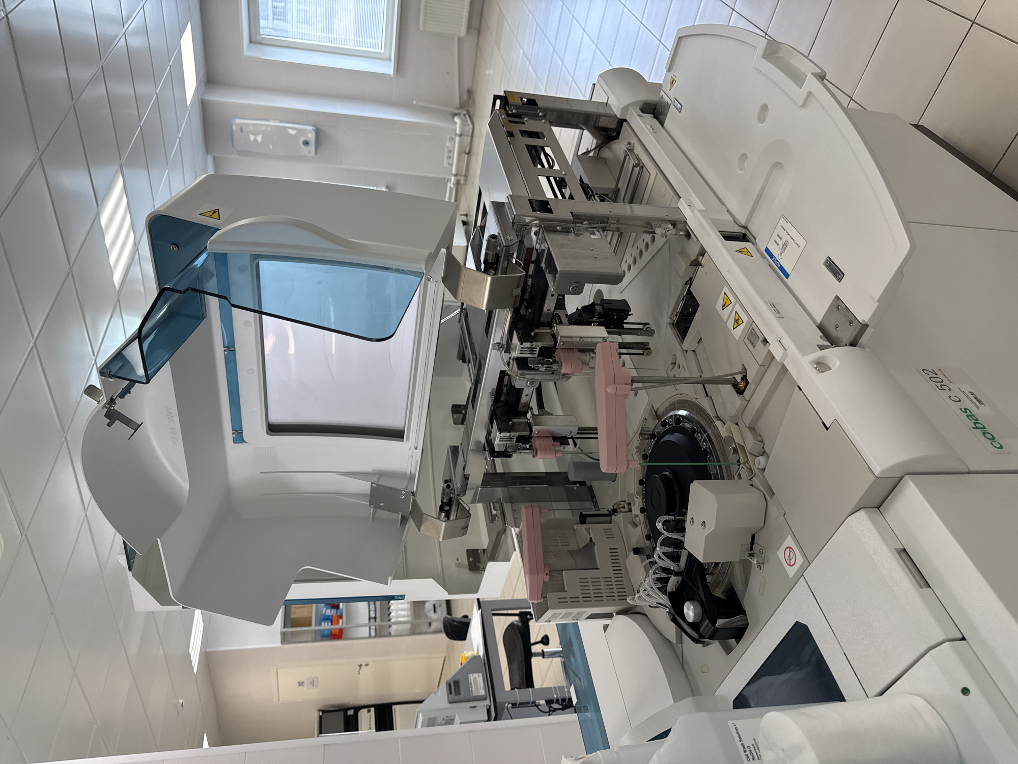 This image depicts a laboratory setting, likely in Moscow, Russia, featuring a prominent automated clinical chemistry analyzer, identified by the label "cobas c 502" on its front panel. The machine is large, predominantly white, and composed of various mechanical and electronic components. Its lid is partially open, revealing an internal mechanism with robotic arms and sample handling systems. Several bottles of reagents are visible to the left of the analyzer, suggesting active use. The surrounding environment appears to be a cleanroom or laboratory space, characterized by tiled floors, a suspended ceiling with fluorescent lighting, and various pieces of laboratory equipment and shelving in the background. The overall impression is one of a sterile, functional, and technologically advanced medical or research facility.
