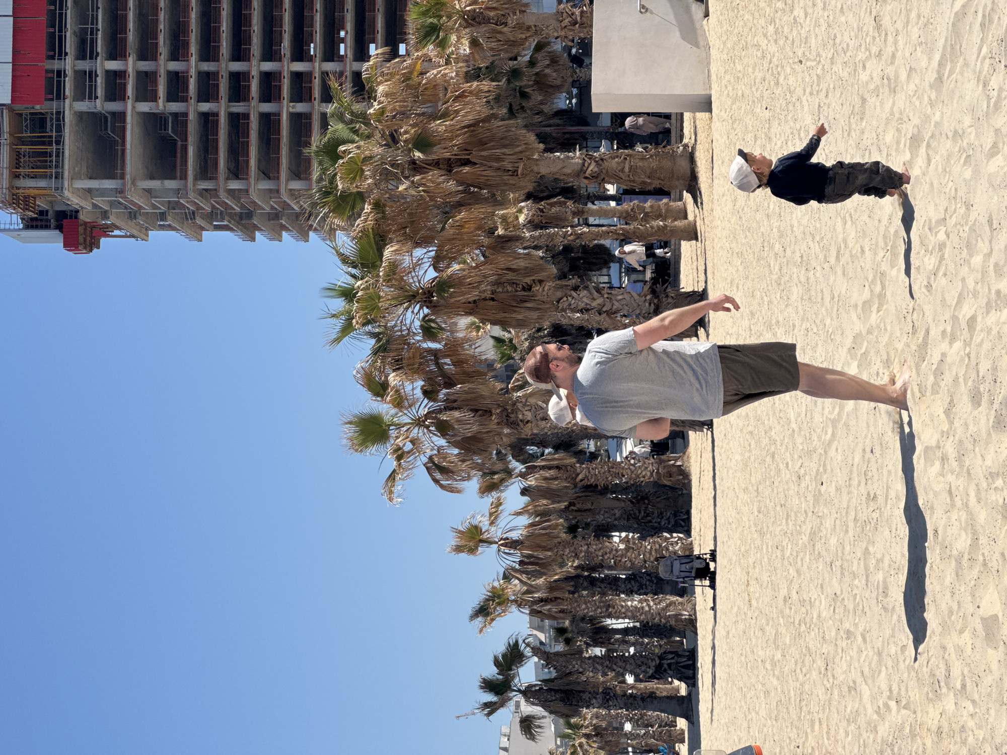 An outdoor, eye-level shot captures a beach scene in Bat Yam, Israel, under a bright, clear blue sky. The image is bathed in strong daylight, casting distinct shadows on the sandy ground, suggesting a sunny day.
In the immediate foreground, a man is visible from the waist down, standing on the light-colored sand. He wears olive-green shorts and a grey t-shirt, with a white baseball cap on his head, facing away from the camera towards the right. Further back and slightly to the right, a young child stands barefoot on the sand, dressed in a dark long-sleeved top, dark pants, and a white baseball cap. The child is actively pointing towards the upper right of the frame, seemingly at something out of view or in the distance.
The midground is dominated by a dense row of palm trees. Many of these trees appear dry or brown, with some patches of green fronds visible, indicating either their natural state or a dry period. Behind the palm trees, in the left background, a large, multi-story building is under construction, its concrete skeleton, exposed rebar, and scaffolding clearly visible. The right side of the background shows more of the clear blue sky and some distant, indistinct structures. The overall setting combines natural beach elements with urban development. There is no visible text in the image.