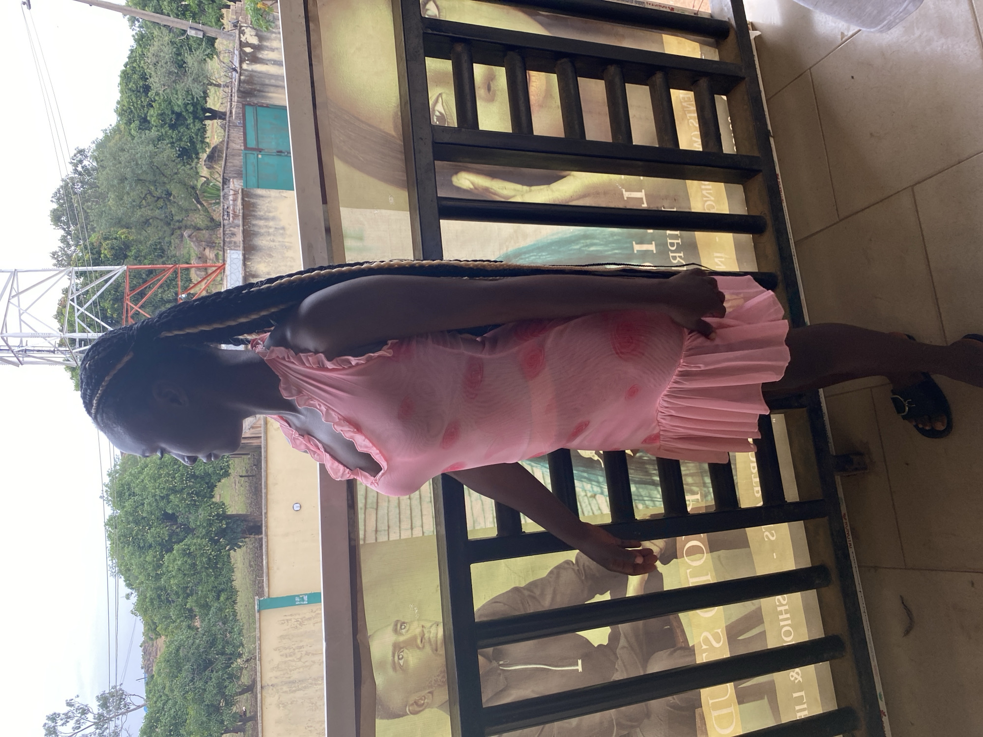 A dark-skinned woman in a pink dress stands behind a black metal railing. The railing overlooks an outdoor scene with trees and a building. A telecommunications tower is visible in the distance. A poster is behind the railing with a portrait of a person. The location is likely Jos, Nigeria, based on the vegetation and the overall setting.