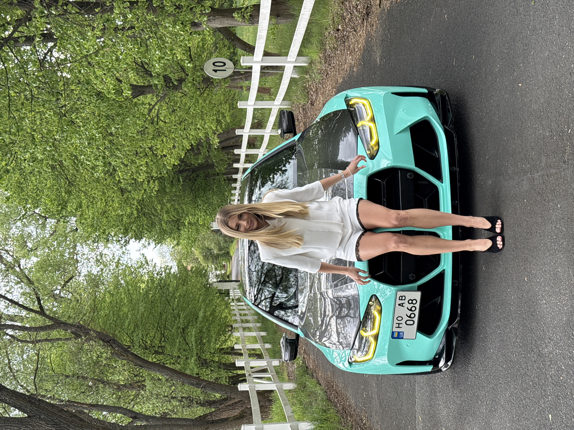 A young blonde woman dressed in white clothing is posing on the hood of a turquoise car with a dark hood. The car has a license plate that reads "HO AB 0668." The scene takes place on a road lined with white fences and green trees. A sign with the number "10" is visible near the road. The location is Unknown, Unknown.