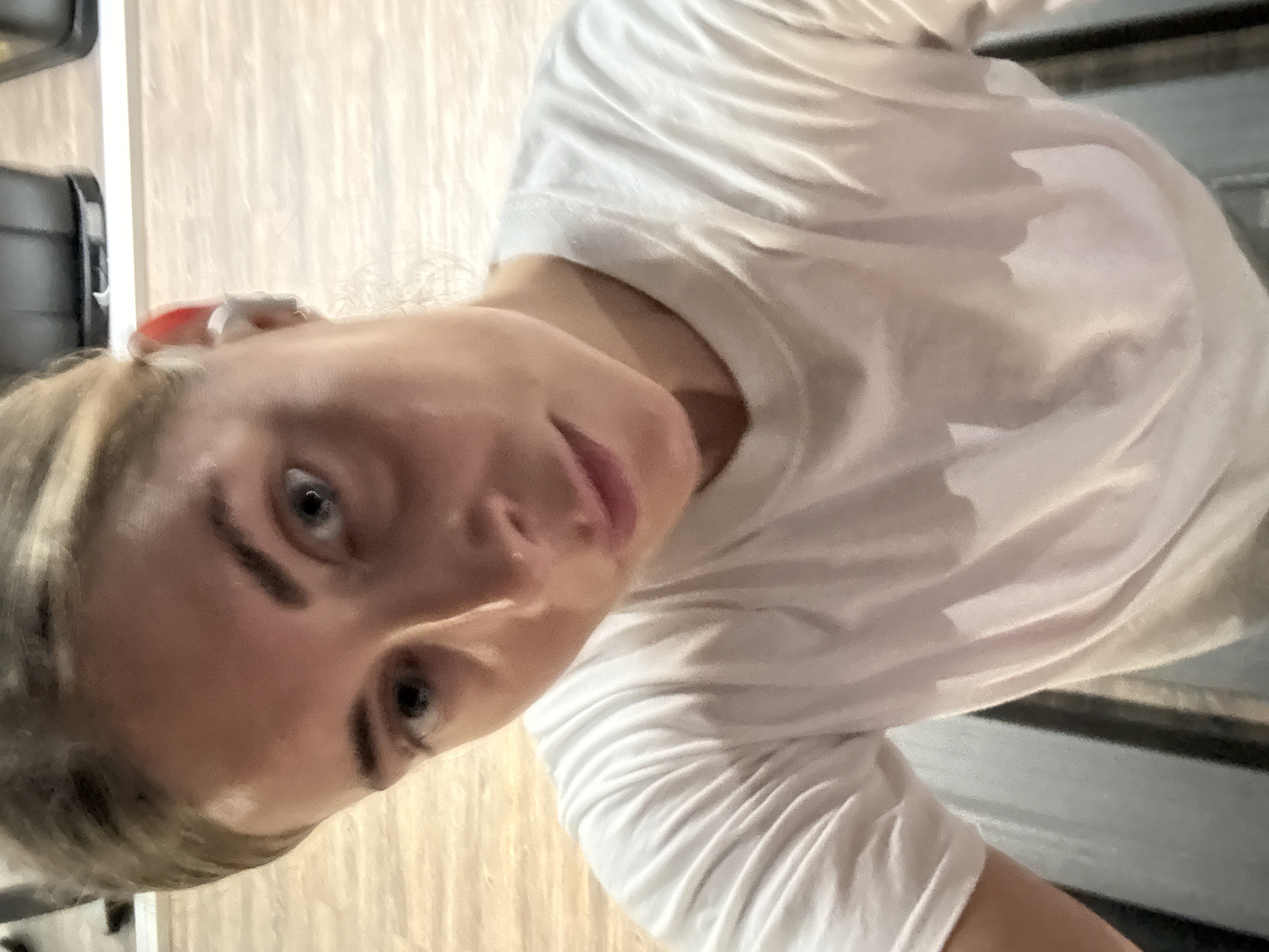 A close-up, slightly angled selfie of a woman with blonde hair and blue eyes wearing a white t-shirt. The background is blurred and appears to be an indoor space with wooden flooring. A piece of what seems to be a treadmill is visible in the upper and lower right corners of the frame. An earbud is in the woman's right ear. The photograph does not provide sufficient information to determine a specific location in Paris, France.