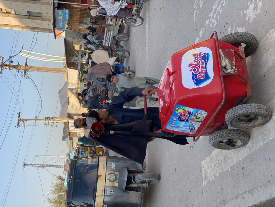 This image captures a busy street scene in Kandahar, Afghanistan, under clear daylight. In the foreground, a young male, dressed in a dark blue traditional garment, pushes a red four-wheeled ice cream cart. The cart features "Payman Ice cream" branding in English and local script, with one label displaying cartoon characters. A black megaphone, draped with black fabric, is attached to a pole carried by the individual. A young boy in a light grey traditional garment walks to the right of the ice cream cart.
To the left, a light-colored, three-wheeled auto-rickshaw (tuk-tuk) is visible, with a passenger inside. Further back, a yellow auto-rickshaw is present. The midground and background show a crowded urban street filled with numerous individuals, many in traditional Afghan attire; some women are seen wearing burqas. Multiple motorcycles, some equipped with cargo beds, are interspersed among the pedestrians, suggesting traffic.
The environment includes numerous utility poles with an extensive network of overhead power lines. Buildings of various architectural styles line the street, including a multi-story light-colored structure on the right and several smaller market stalls or shops with awnings. A shop sign with partial Dari/Pashto script is visible. Distant mountains are discernible against the light blue sky. Solar panels are observed on some rooftops. The ground surface is an unpaved or dirt road.