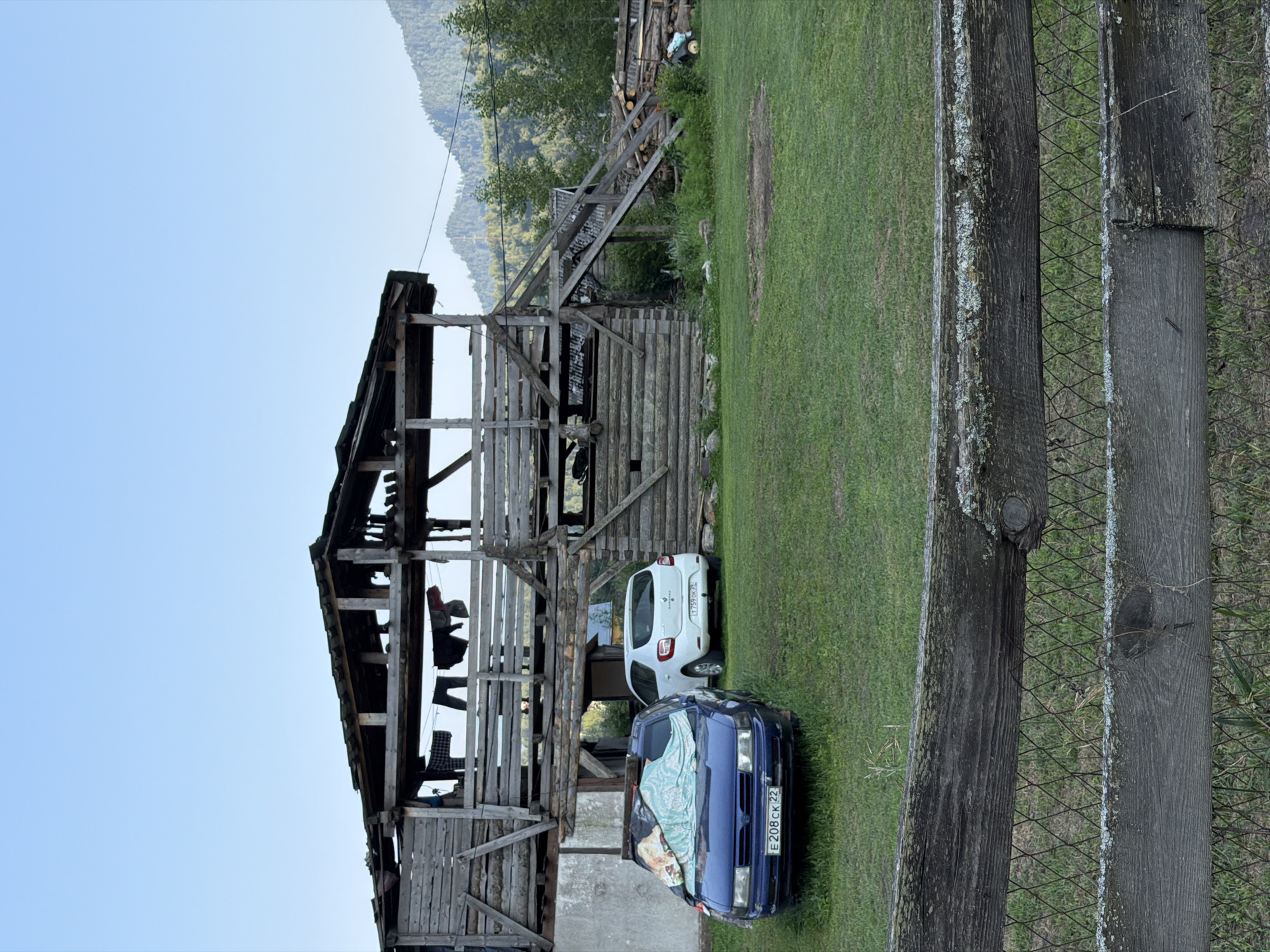 The image depicts a rural scene in the vicinity of Anos, Russia, with a focus on a rustic wooden structure and vehicles. The foreground features a weathered wooden fence with wire mesh, set against a backdrop of green grass. Behind the fence, two cars are parked on the grass. The car on the left is blue and covered with a light-colored tarp, while the car on the right is white.
Dominating the midground and background is a substantial, multi-level wooden structure, appearing to be a barn or storage building. It is constructed from rough-hewn logs and planks, with open sides and a sloped roof, some sections of which appear damaged or incomplete. Laundry is hanging on a line across the upper level of the structure. To the right of the wooden building, a pile of logs is visible, along with some gardening tools.
The environment suggests a mountainous or hilly region, with a forested mountainside visible in the far background under a clear blue sky. The overall impression is one of a functional, perhaps aged, rural homestead in a natural setting. No people are visible in the frame.