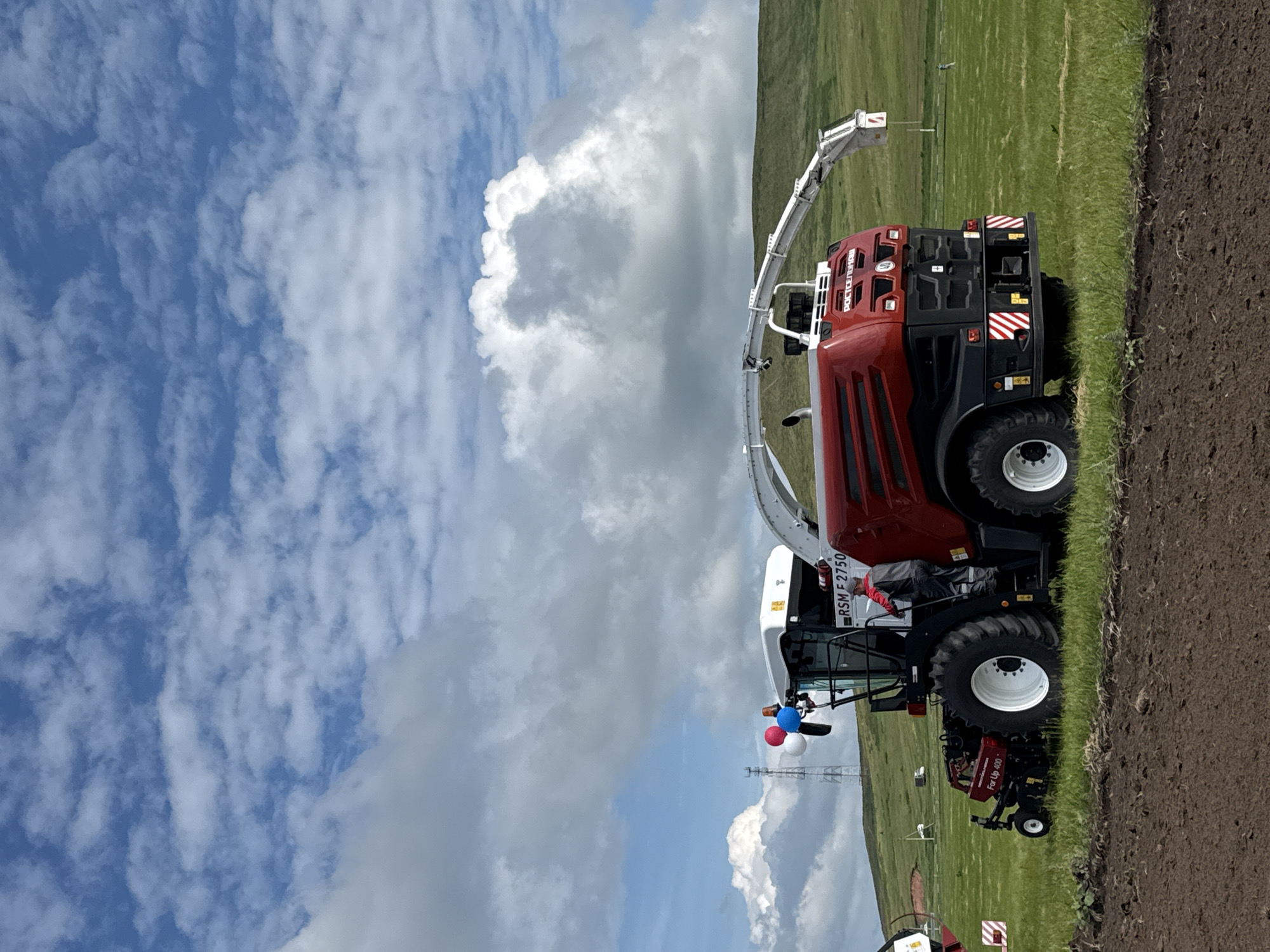 An agricultural combine harvester, predominantly red with black accents and a white cab, is parked in a grassy field under a partly cloudy sky. The combine has large rear wheels and a smaller front wheel assembly with a piece of agricultural equipment attached. A person wearing a gray uniform stands in the open doorway of the combine's cab. Three balloons, red, white, and blue, are attached to the top of the cab. In the background, rolling green hills are visible. The foreground shows freshly tilled dark brown soil. A cell tower stands in the distance to the left of the combine. The scene appears to be an outdoor event or demonstration, possibly in the city of Nurkeevo, Russia.