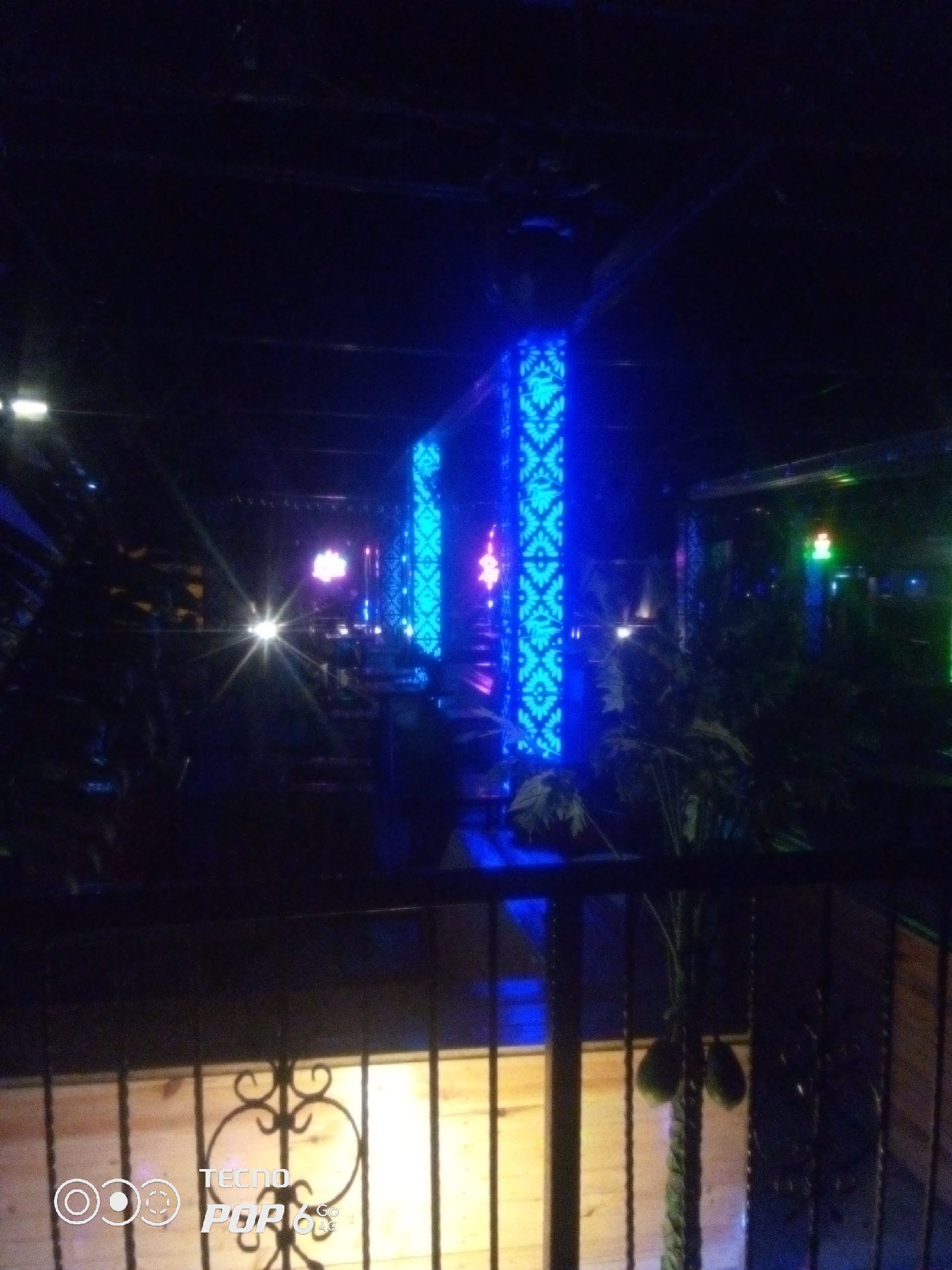 This is a nighttime photograph taken from an elevated position, looking down into an indoor space with a modern, dimly lit ambiance. The scene is dominated by a prominent row of tall, rectangular pillars illuminated with intricate blue patterns, creating a striking visual effect. The pillars appear to be part of a structural element or decorative feature within the venue.
In the background, various lights are visible, including what appears to be a bright starburst light source in the center, possibly from a spotlight or a lens flare. There are also hints of other colored lights, such as pink and green, suggesting a dynamic lighting setup. Plants, possibly palm trees, are interspersed throughout the space, adding a touch of greenery to the artificial lighting.
The foreground features a dark metal railing with decorative scrollwork, indicating a balcony or a viewing platform. Below the railing, a wooden floor is visible, illuminated by ambient light. A watermark in the bottom left corner of the image reads "TECNO POP 6 4G".
The overall impression is of a nightclub, lounge, or a similar entertainment venue. The lighting and modern design suggest a place that is active in the evening. There are no people clearly visible in the image, and no specific actions or interactions can be discerned. The location is identified as Howayi, Nigeria, suggesting this is a popular spot in that city.