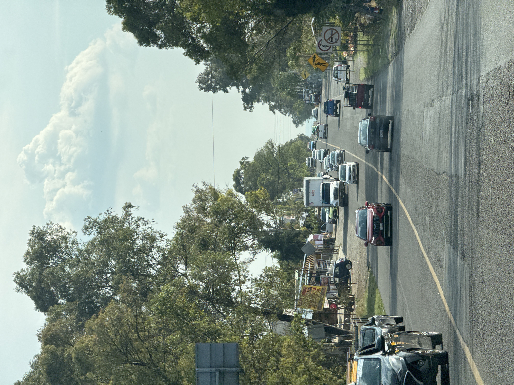 The image, correctly oriented, displays a daytime scene of a two-lane asphalt road in San Pablo Ahuatempa, Mexico, experiencing moderate to heavy traffic. Numerous vehicles, including sedans, SUVs, pickup trucks, and a white commercial truck, are visible in a line, suggesting slow-moving traffic or a queue primarily on the right side of the road.
The road features a yellow double center line and a white shoulder line. Both sides of the road are lined with abundant green trees, some forming a dense canopy. On the left side, behind the trees, several informal structures, likely market stalls or small shops, are discernible, adorned with various banners and signs.
The sky is bright and partly cloudy, with large, fluffy white cumulus clouds prominent on the left side, indicating fair weather. Shadows cast by vehicles and trees confirm it is daytime.
Notable details include several road signs on the right side of the road. A yellow diamond-shaped sign features a black pedestrian crossing symbol. Below it are two white circular signs with red diagonal lines, one indicating "no parking" and the other possibly "no passing." On the side of the white commercial truck, the letters "ABG" are faintly visible. Among the signs for the stalls on the left, fragmented text such as "MA NA" and "TACO" can be discerned on a yellow banner. The scene captures a typical bustling roadside environment.