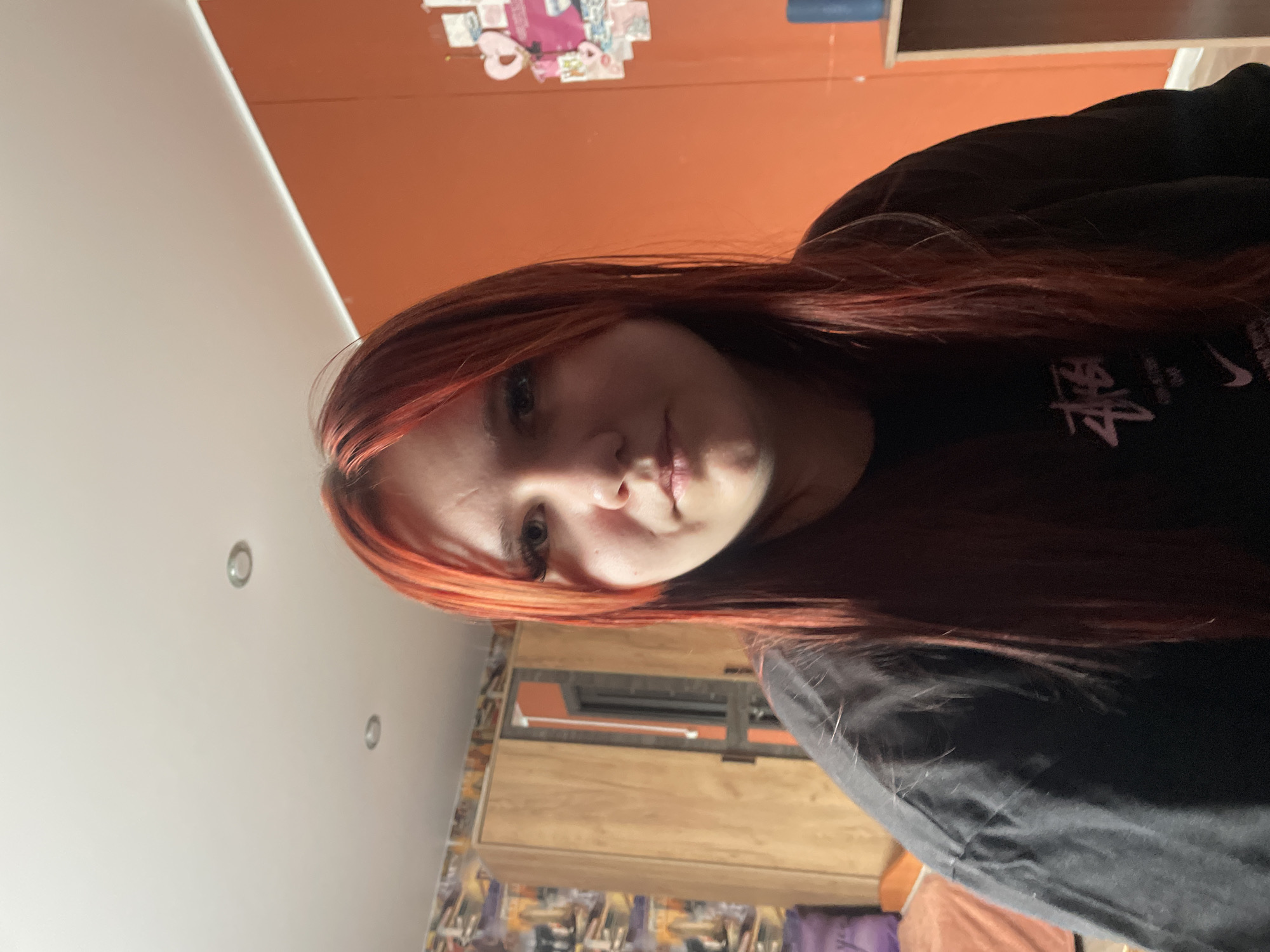The image is a selfie taken indoors, likely in a bedroom in the city of Медведево, Russia. A young woman with long, reddish-orange hair and bangs is the central subject, looking directly at the camera. She is wearing a black t-shirt with a graphic logo on it. The background shows a wooden wardrobe with a mirror, an orange wall adorned with various pictures and stickers, and a ceiling with two circular lights. Part of a bed with a purple pillow and an orange blanket is visible on the left.