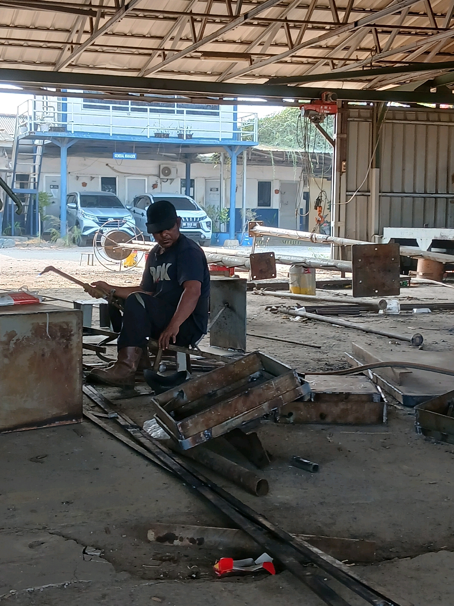 The image depicts an outdoor industrial workshop located in Pangkalpinang, Indonesia. A man, wearing a black t-shirt, black pants, a black cap, and brown work boots, is seated on a stool in the center of the frame. He is holding a metal tool in his right hand and appears to be working on a metal object. The workshop is filled with various metal materials, including pipes, sheets, and what appear to be molds. In the background, there are two cars parked in front of a two-story building with blue accents and a sign that reads "GENERAL MANAGER." A red overhead crane is visible on the left side of the background. The ground is a rough, concrete or dirt surface, scattered with debris. The overall environment suggests a busy, active worksite.