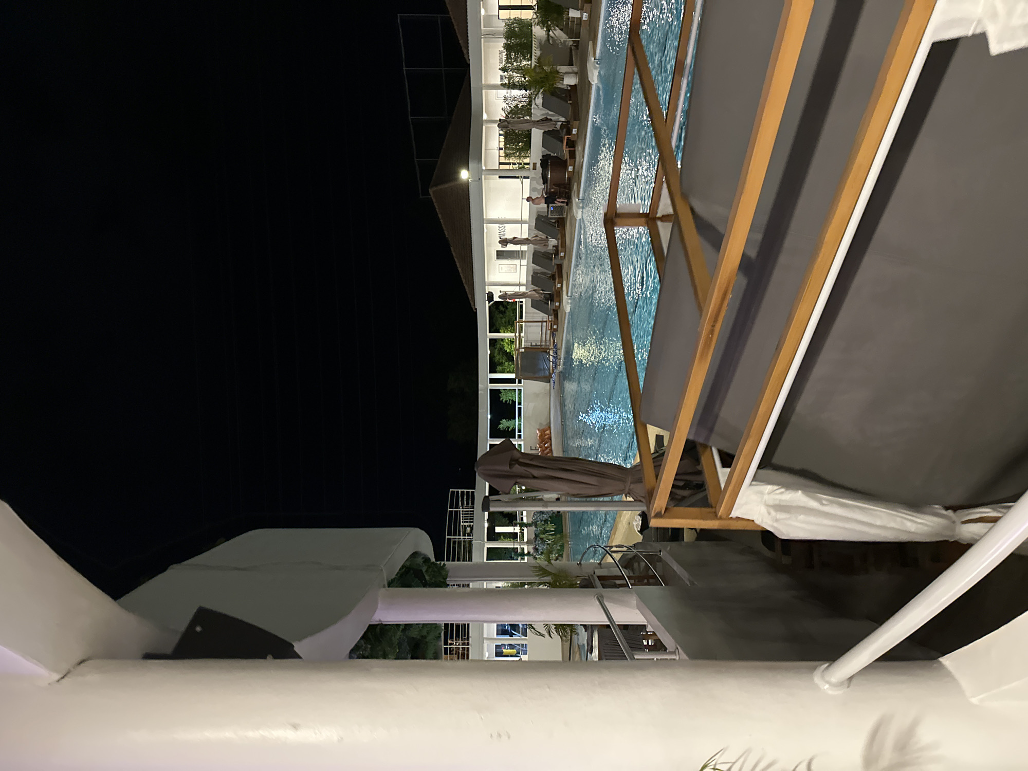 A night-time, eye-level shot captures a swimming pool area at a resort. In the foreground, a white pillar and railing are visible to the left, with a dark grey canopy structure supported by wooden beams to the right. The swimming pool, illuminated by overhead lights, is the central feature, with blue water reflecting the light. Lounge chairs and umbrellas are arranged around the pool. In the background, a building with a white facade and a brown roof is visible, with lights on inside. Several palm trees are scattered throughout the scene. The sky is dark and clear. There are no discernible people in the image, other than a distant figure near a hot tub. The location is identified as Unknown, Unknown.
