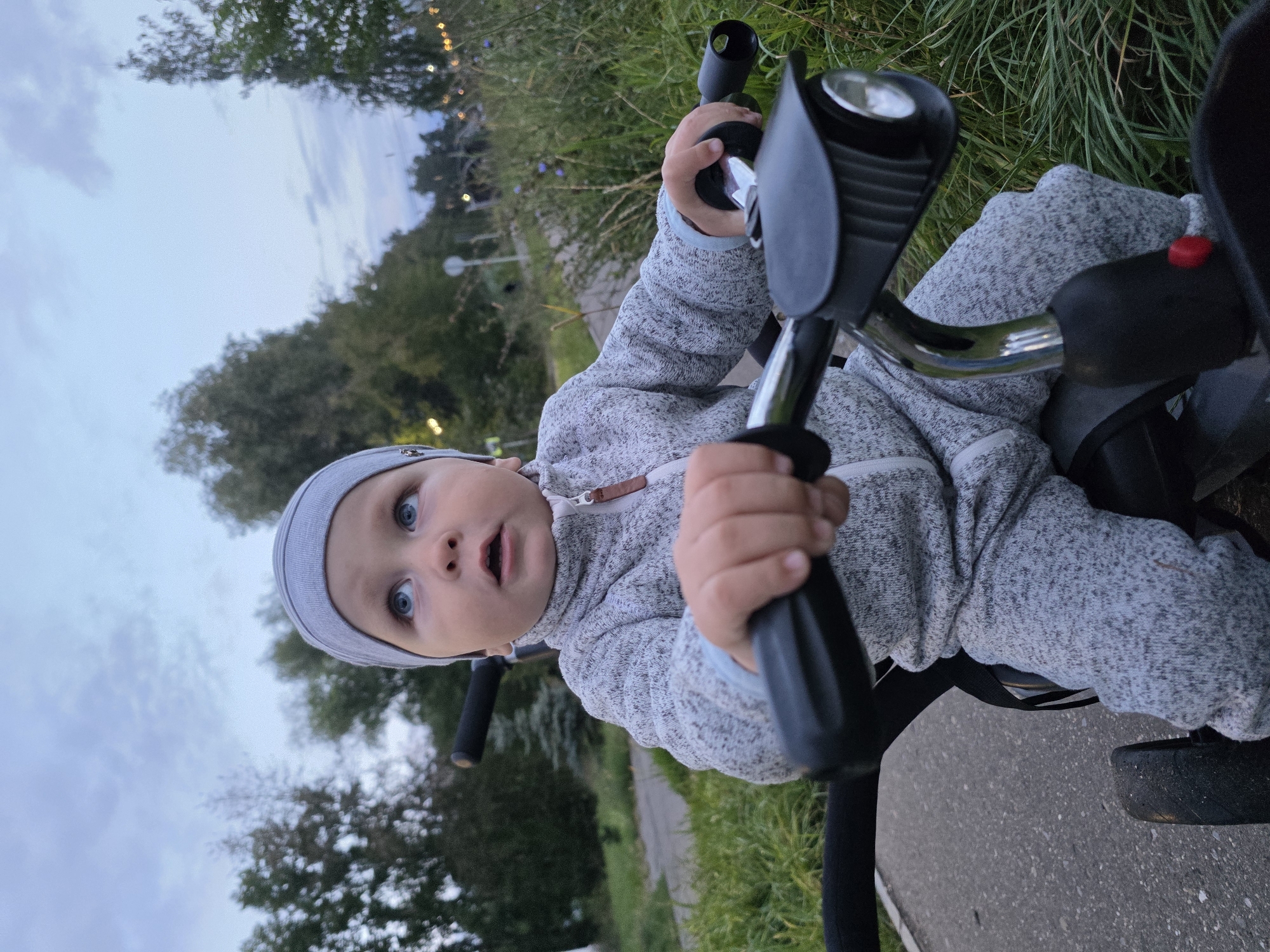 The image shows a young child wearing a grey beanie and a grey textured tracksuit, sitting on a black tricycle with handlebars. The child is looking up and to the right with a curious expression. The tricycle has a black headlight attached to the handlebars. The scene takes place outdoors on a paved path, with a grassy area and trees in the background. The sky is cloudy, suggesting it is either early morning or late evening. The presence of string lights visible in the background suggests this location may be a park or a public area in the city of Chaikovsky, Russia, possibly decorated for an event.