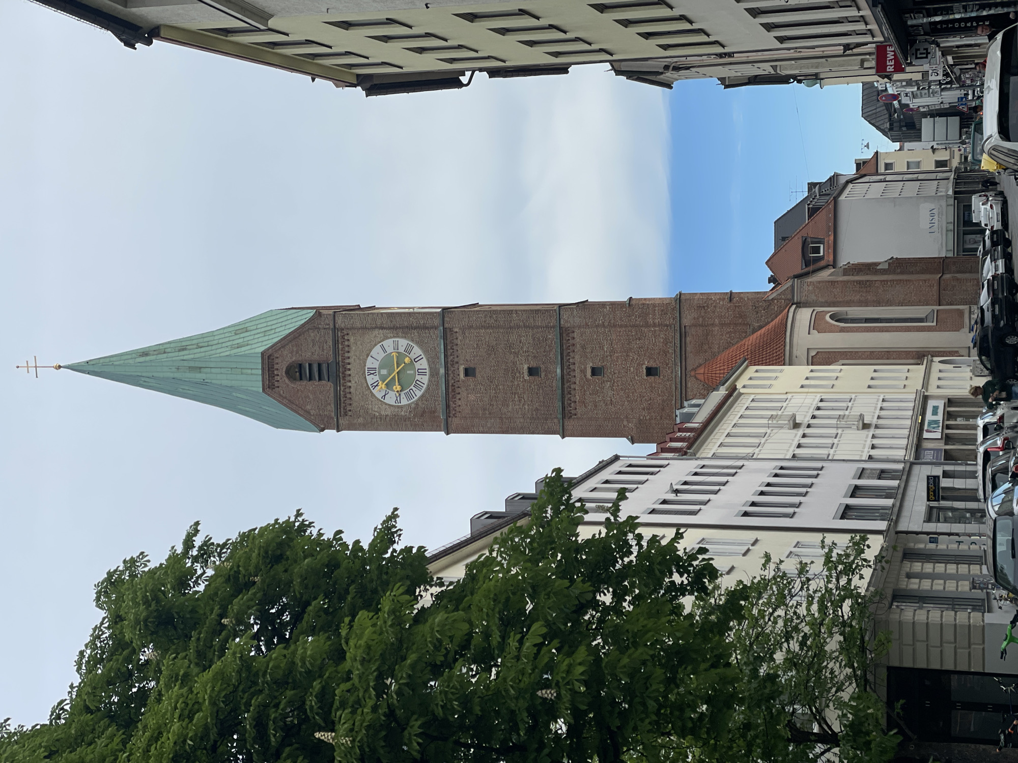 The image captures a view looking down a narrow urban street lined with multi-story buildings, dominated by a tall church tower on the left. The prominent church tower is constructed of reddish-brown brick, featuring a distinctive green, pointed spire topped with a golden cross. A large clock face with Roman numerals adorns the tower, with hands indicating approximately 10:11 or 10:12.
In the foreground, lush green trees with dense foliage partially frame the bottom of the scene, obscuring some of the buildings. On both sides of the street, various buildings display a mix of architectural styles, some with numerous rectangular windows and different rooflines.
Further down the street, on the right side, multiple cars are parked along the curb. A few indistinct figures, likely pedestrians, are visible in the distance. The sky above is a mix of light blue patches and soft white clouds, suggesting a bright, partially overcast daytime.
Visible text includes "REWE" on a red sign, "UNISON" on a white building, "gangbild" on a yellow sign, and "ATELIER" on a grey sign. Another sign with "LIZON" or "LISON" is visible on a building further down the street. The scene depicts a typical urban day, with no specific event or activity taking place beyond general street life.