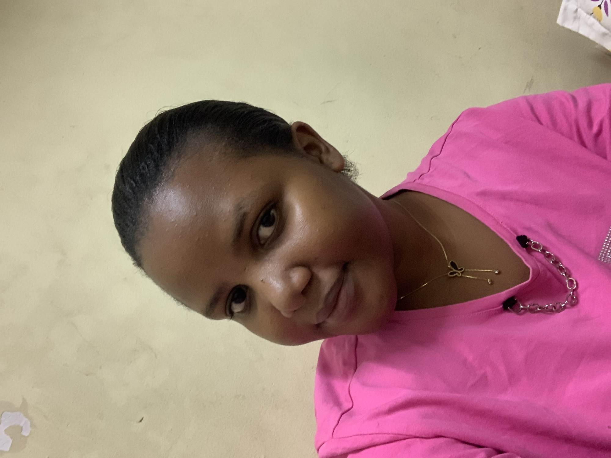 A woman with dark hair, brown eyes, and dark skin is wearing a pink top. She is smiling slightly and looking directly at the camera. She is wearing a gold necklace with a butterfly pendant and a silver chain. The background is a plain, pale yellow wall. The location appears to be indoors, but no further details indicate that this is in Dar es-Salaam, Tanzania.