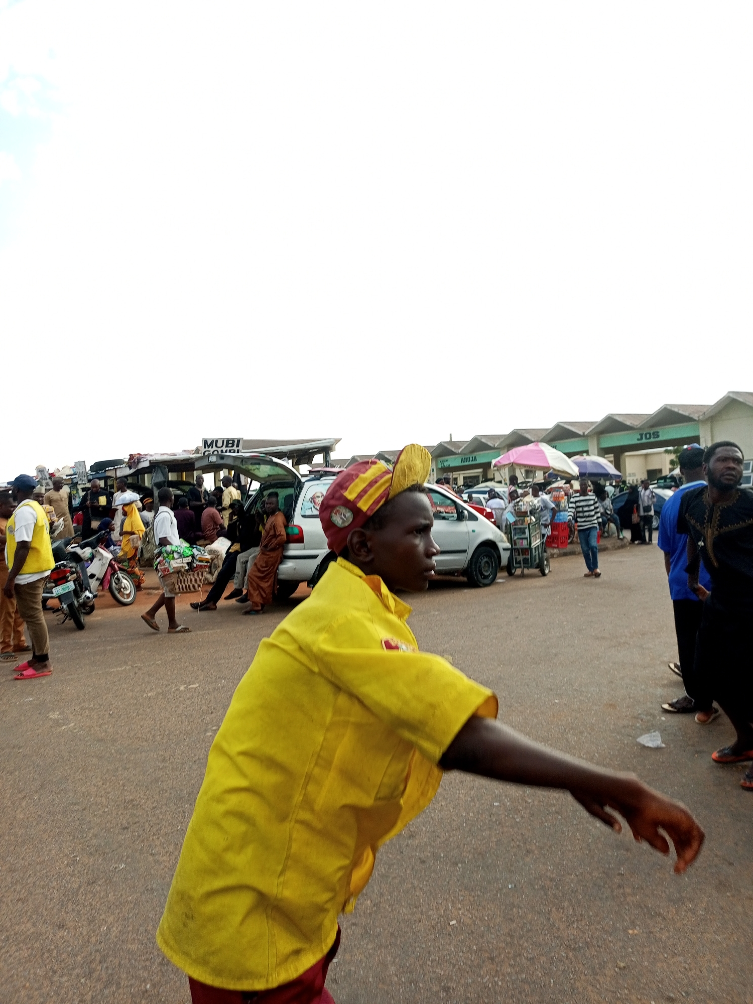 The image depicts an outdoor scene in the city of Gombe, Nigeria, likely a market or transportation hub. In the foreground, a young person in a bright yellow shirt and a red and yellow cap is walking, their body angled towards the right of the frame. The person's arm is extended, suggesting they might be gesturing or reaching. Behind them, a silver vehicle with its rear hatch open is parked on a paved surface, with several people seated or standing around it. Other vehicles, including motorcycles, are visible in the background. Buildings with signs, one reading "MUBI CONDI" and another with "JOS," line the area, suggesting commercial activity. People of various ages and genders are present, some carrying goods or interacting with each other. The sky is overcast and bright. The overall atmosphere appears busy and typical of a commercial district.