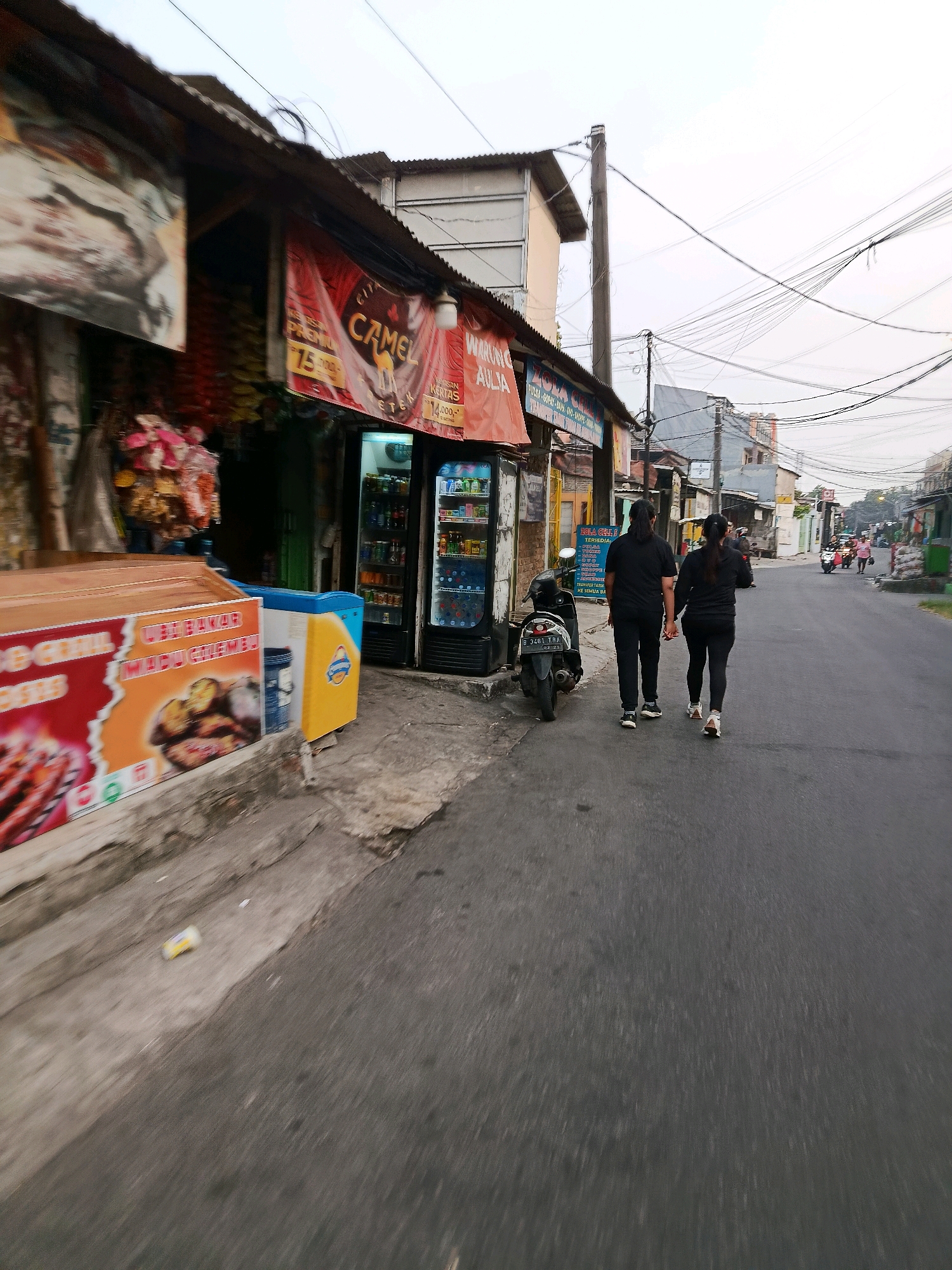 The image depicts a street scene in Bekasi, Indonesia. Two individuals, one with long dark hair and the other with shorter dark hair, both wearing black athletic wear, are walking away from the camera down a paved road. They are holding hands. To their left, a row of small shops lines the street. One shop has a red banner with "CAMEL" prominently displayed. In front of the shops, there is a motorcycle parked. Further down the street, other buildings and a few more motorcycles are visible. The sky is overcast, and overhead power lines crisscross the scene. The overall impression is of a typical street in a residential or commercial area of Bekasi.