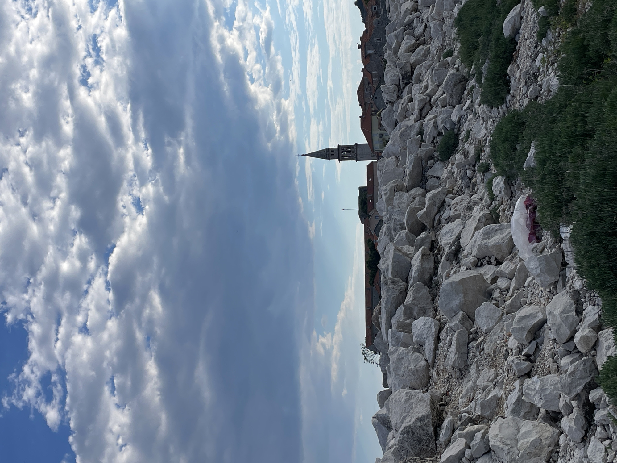 The image presents a view from a rocky slope towards a city skyline under a partly cloudy sky. The foreground is dominated by light-colored rocks and sparse vegetation. In the middle ground, a tall church tower with a pointed spire stands out against the sky. Adjacent to the tower are buildings with red-tiled roofs, suggesting an urban environment. The sky is filled with a mix of puffy white clouds and blue patches. The location is unknown.