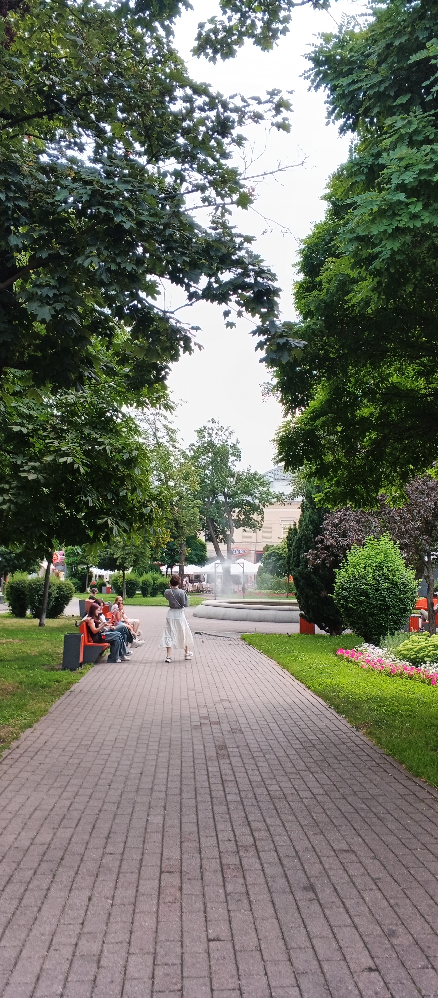 This is a vertical, medium-angle, outdoor shot of a paved walkway in a park-like setting. In the foreground, the walkway is constructed of grey brick pavers arranged in a diagonal pattern. To the left of the walkway is a grassy area with a line of orange and grey park benches occupied by at least four individuals. Further into the park, a woman in a light-colored skirt and grey top is walking away from the camera down the walkway. In the mid-ground, a circular fountain with mist spraying upwards is visible. Behind the fountain, there are buildings with awnings and a sign for "Moya", suggesting a commercial area adjacent to the park. The background is dominated by mature trees with dense green foliage, and the sky is overcast. The scene suggests a public urban space, possibly a plaza or park in Kyiv, Ukraine, given the presence of the building signage.