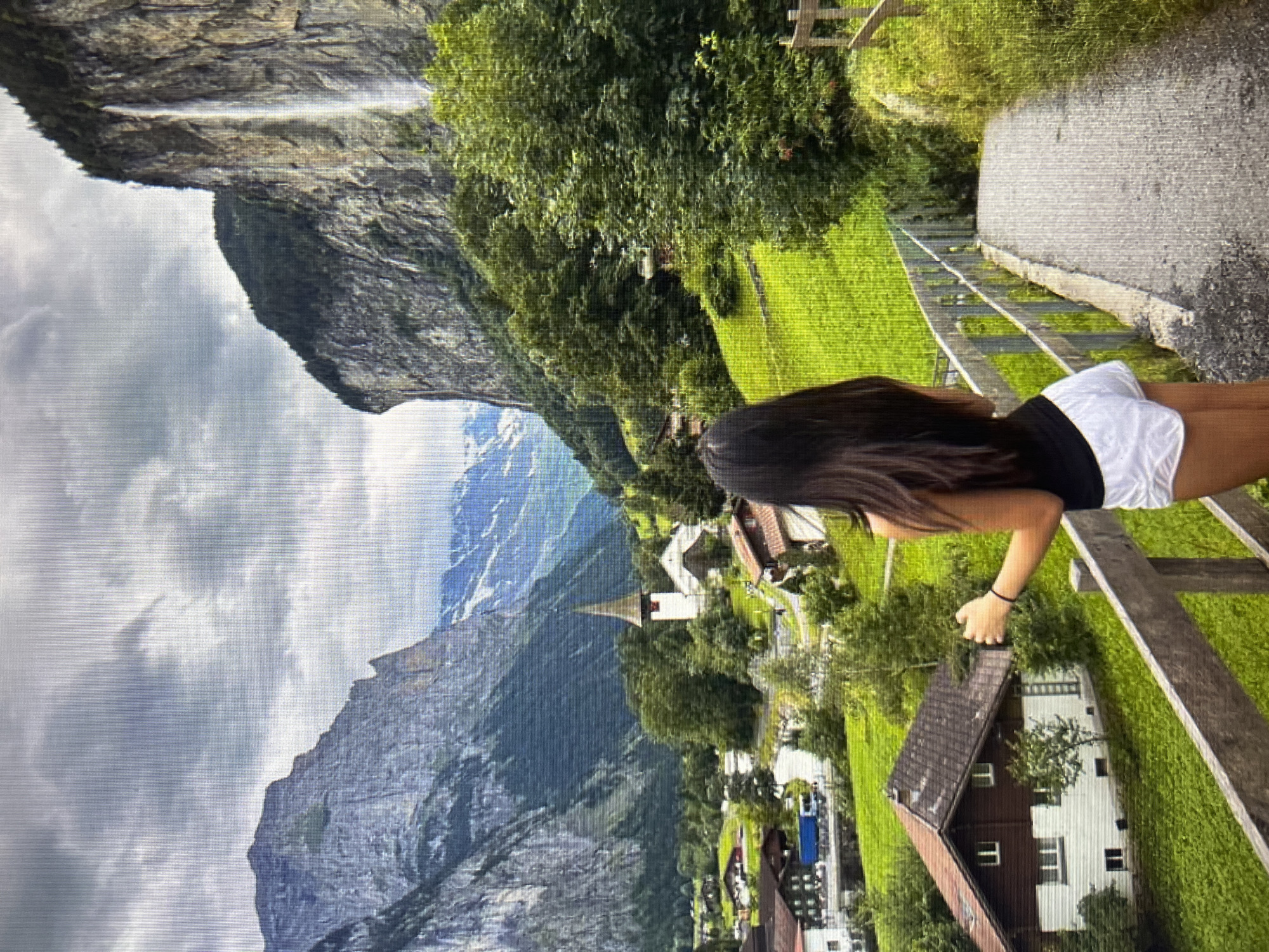 An overcast, cloudy sky looms over a picturesque valley with steep, rocky mountains on either side. A thin waterfall cascades down the right-hand cliff face. In the foreground, a person with long, dark hair, wearing a black tank top and white shorts, stands on a gravel path, facing away from the camera and looking out at the scenery. A wooden railing lines the path. The valley floor is a vibrant green, dotted with charming houses with red roofs and a white church with a steeple. A blue truck is visible on a road winding through the village. The overall scene depicts a serene mountain village landscape.