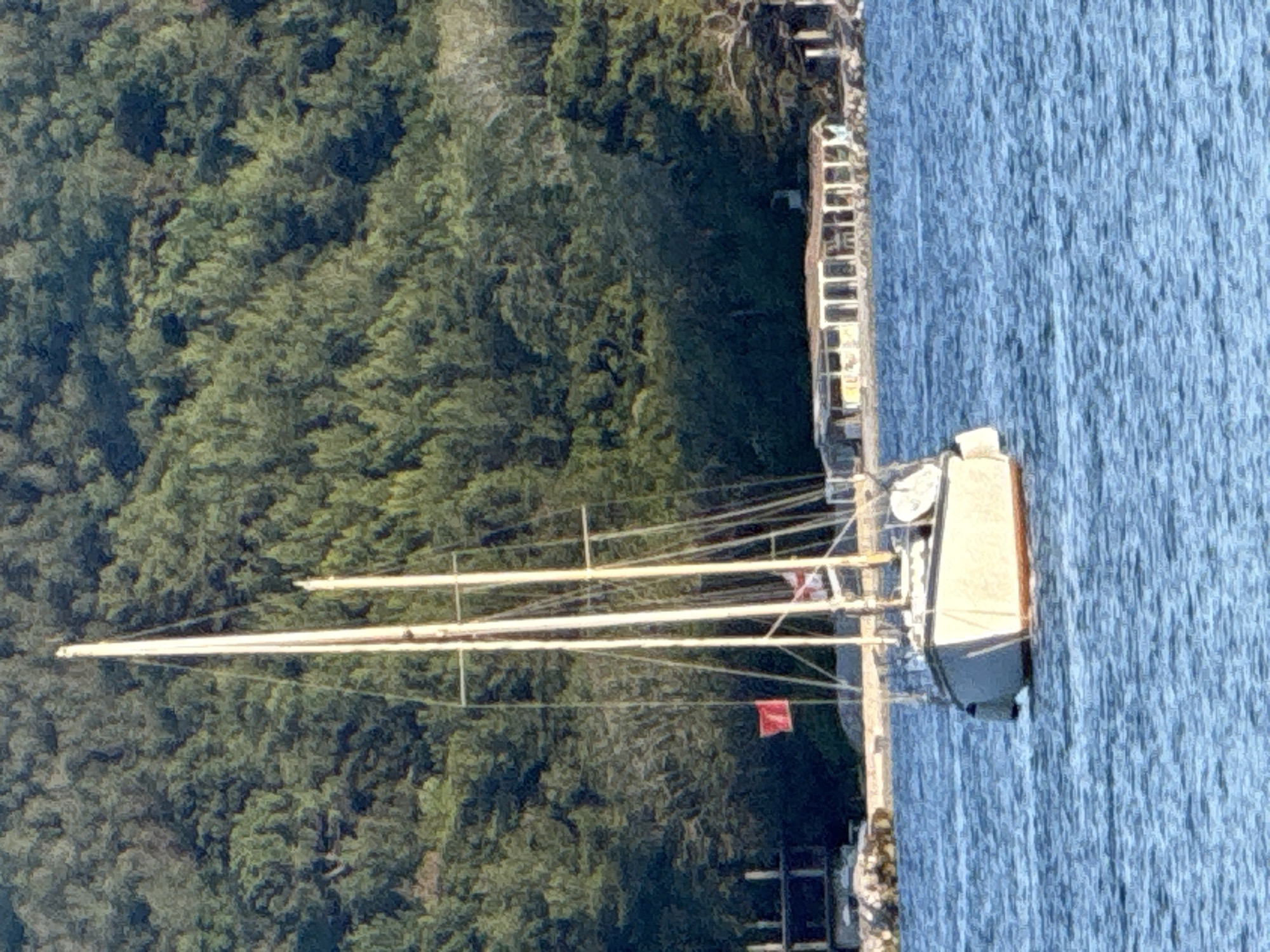 A sailboat with two masts is moored in the water, with a lush green forested hill in the background. A small red flag is visible on the forward mast. On the shore, there is a long, low building with a covered patio area, and several other structures. The water is blue and appears to be slightly choppy. The overall scene is peaceful and idyllic.