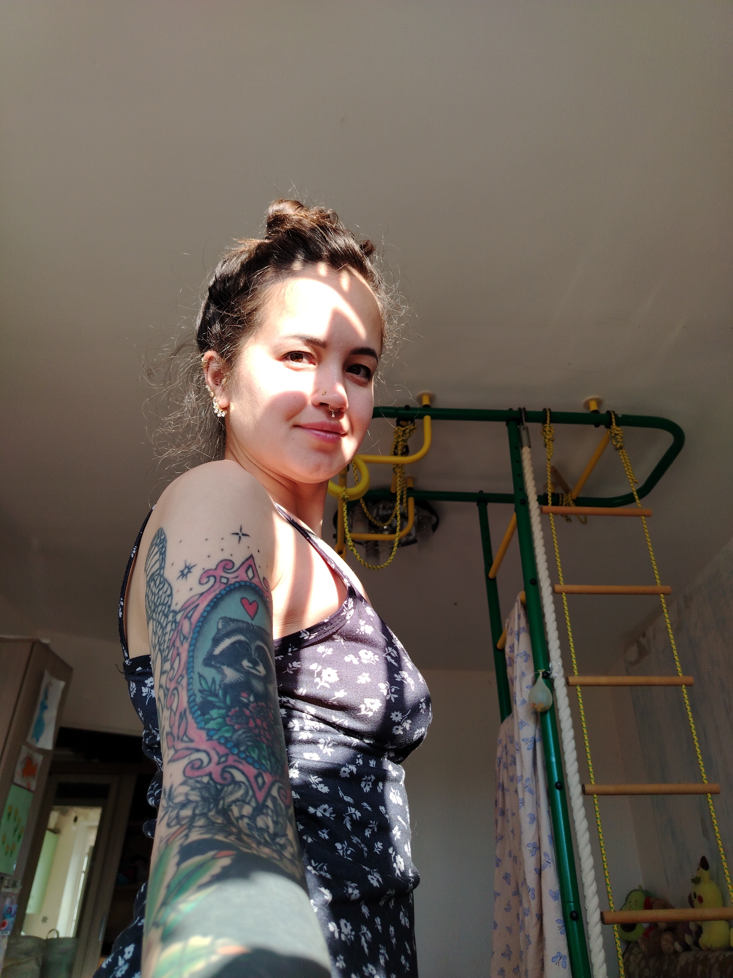 A young woman with dark hair styled in a bun, wearing a dark floral print dress, is shown in a close-up, medium-low angle shot. She has a nose ring and an earring. Her left arm is heavily tattooed, featuring a prominent raccoon tattoo within a decorative frame. Sunlight is casting shadows across her face and body. In the background, a green and yellow indoor gym structure with a ladder made of wooden rungs and rope is visible. The environment appears to be an indoor residential space. The specific location within the city of Noviy, Russia, cannot be determined from the provided image.