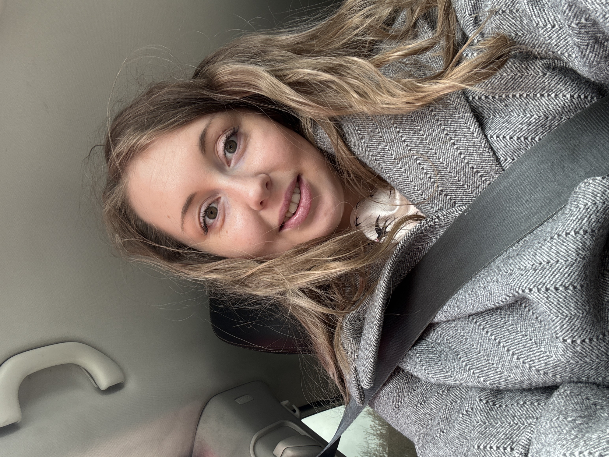 A woman with long, blonde hair is in a car, wearing a herringbone pattern coat and a seatbelt. The car interior is light grey with a white grab handle visible. The location is Unknown, Unknown.