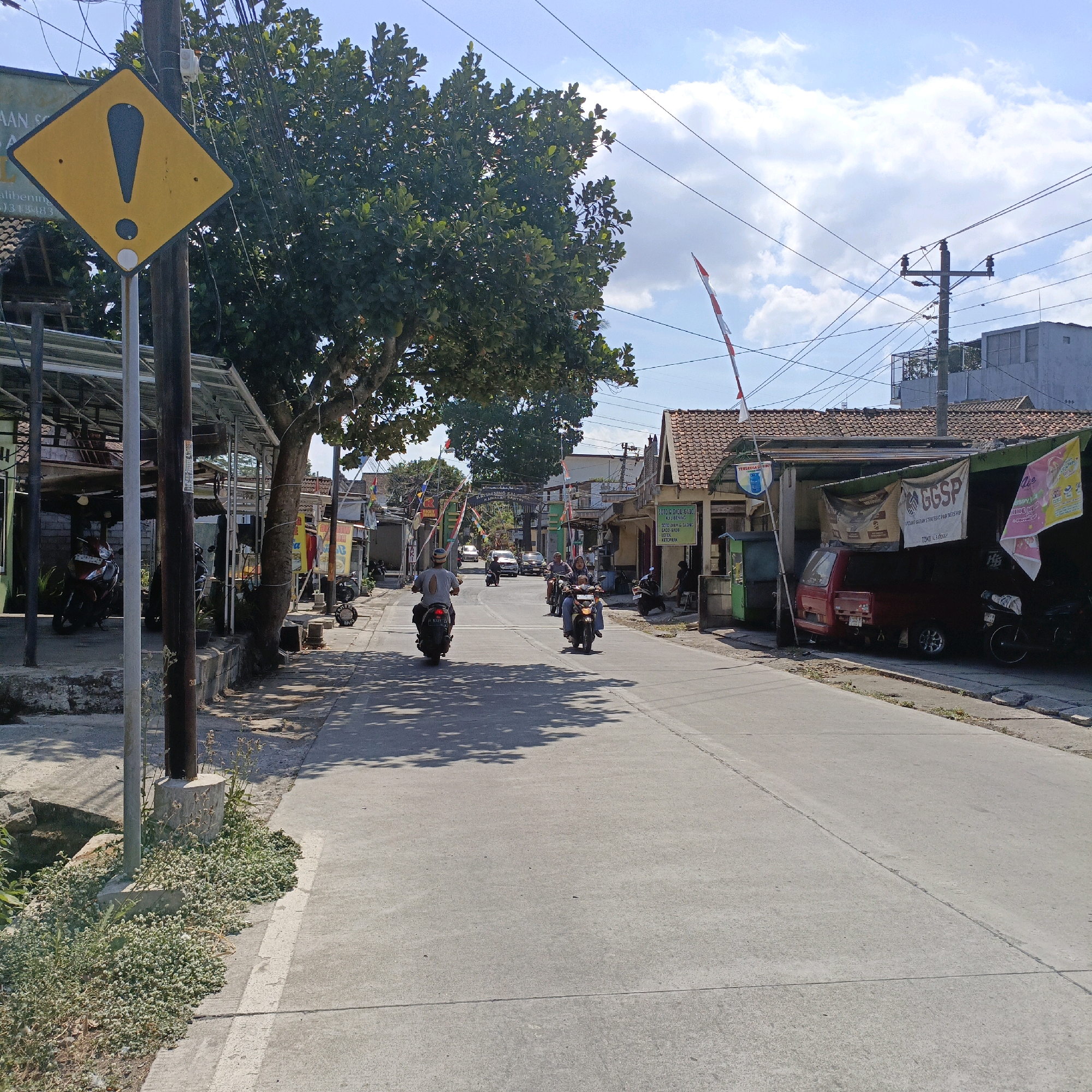The image depicts a street scene in Salatiga, Indonesia, under a bright, cloudy sky. A yellow diamond-shaped sign with a black exclamation mark is prominently displayed on a pole to the left, cautioning drivers. The street is paved with asphalt and lined with buildings on both sides, many of which appear to be shops or small businesses with awnings and signage.
Several motorcycles are visible, with some riders actively traveling along the road. In the foreground, a motorcycle with two riders is moving away from the camera, casting a shadow on the asphalt. Another motorcycle with a single rider is ahead and to the left. Further down the street, more motorcycles and a few cars are present, indicating moderate traffic.
The environment includes a large tree with dense foliage casting shadows on the street. Overhead, electrical wires crisscross the sky, supported by utility poles. Small red and white Indonesian flags are also seen mounted on poles along the street, possibly indicating a national celebration or event. Vegetation, including grass and small plants, grows along the edge of the road and around the base of the utility pole. One building on the right side has a red pick-up truck parked underneath its awning, alongside other motorcycles and banners advertising goods or services. The overall impression is that of a typical bustling street in a town or city.